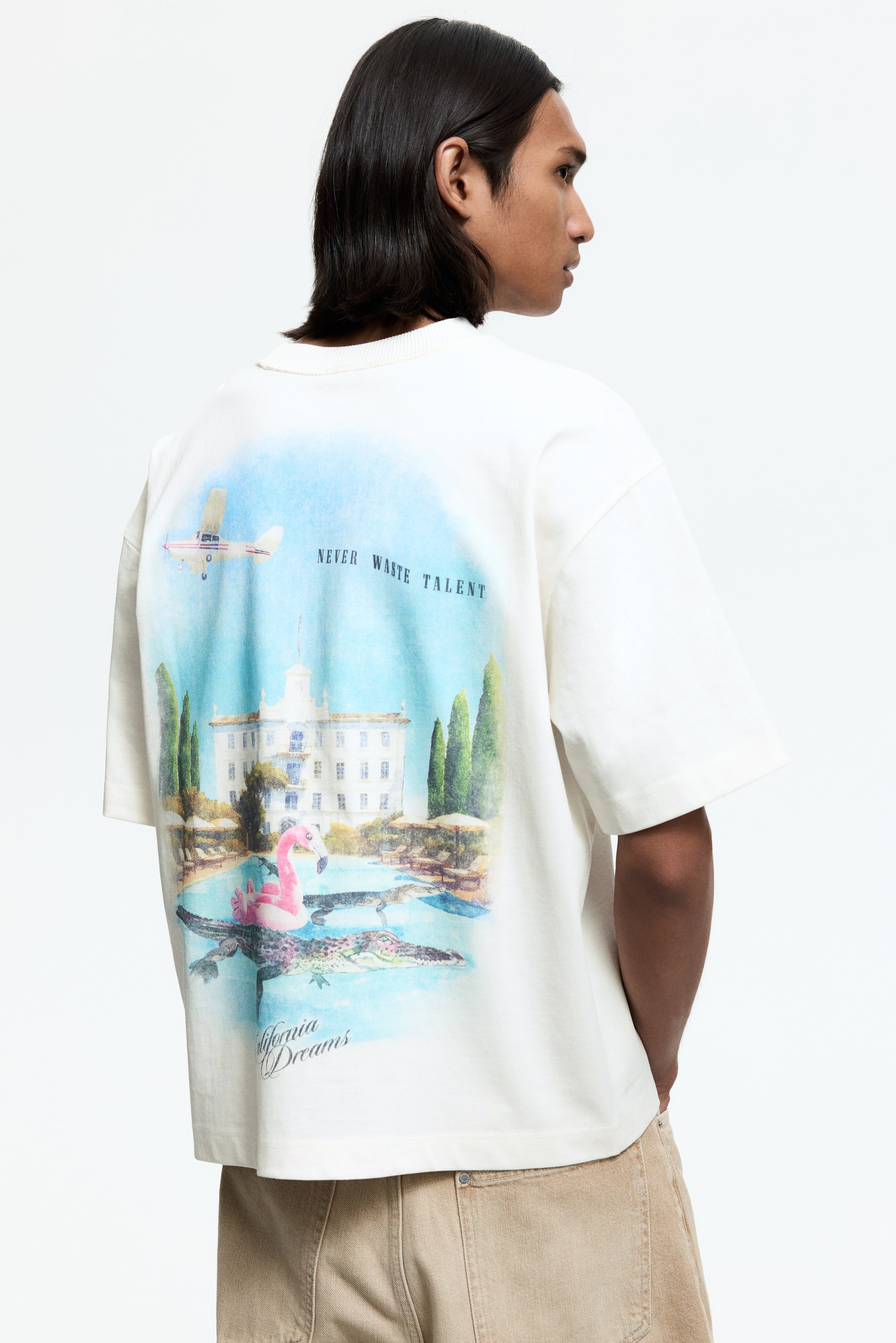 View larger image: The reverse side of an off-white short-sleeve top is showcased, featuring a ribbed crew neckline. A large graphic print adorns the back, illustrating a mansion with a pool, a pink flamingo float, crocodiles, an airplane, and the phrases 'never waste talent' and 'California dreams'.