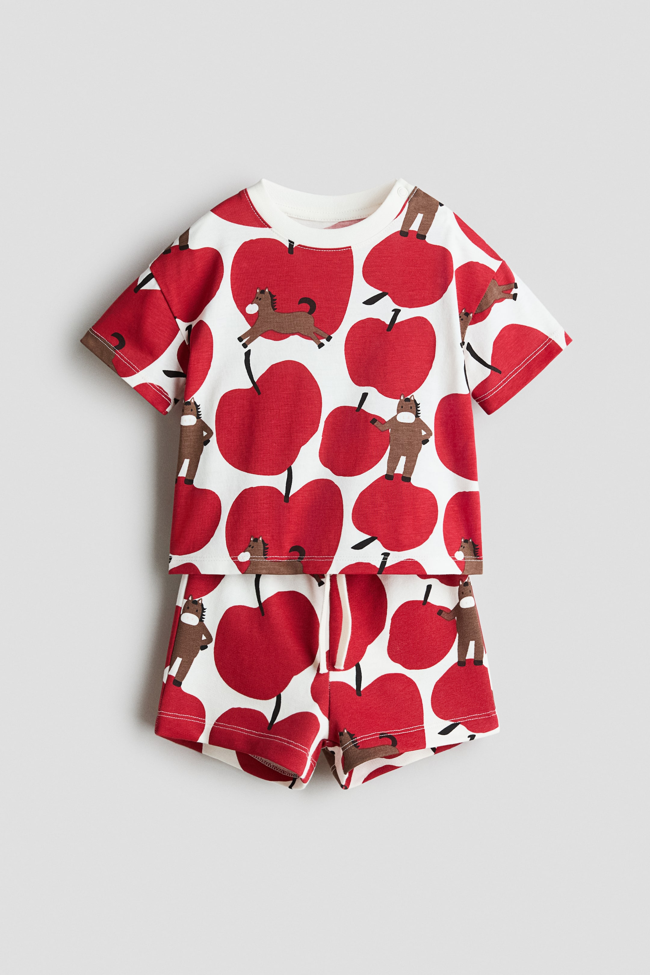 View larger image: A two-piece infant set is including a short-sleeved t-shirt and matching shorts, both facing front. The set is displaying an all-over pattern of red apples and brown horses on a white background, and the t-shirt is designed with a ribbed crew neck and snap buttons on the left shoulder.