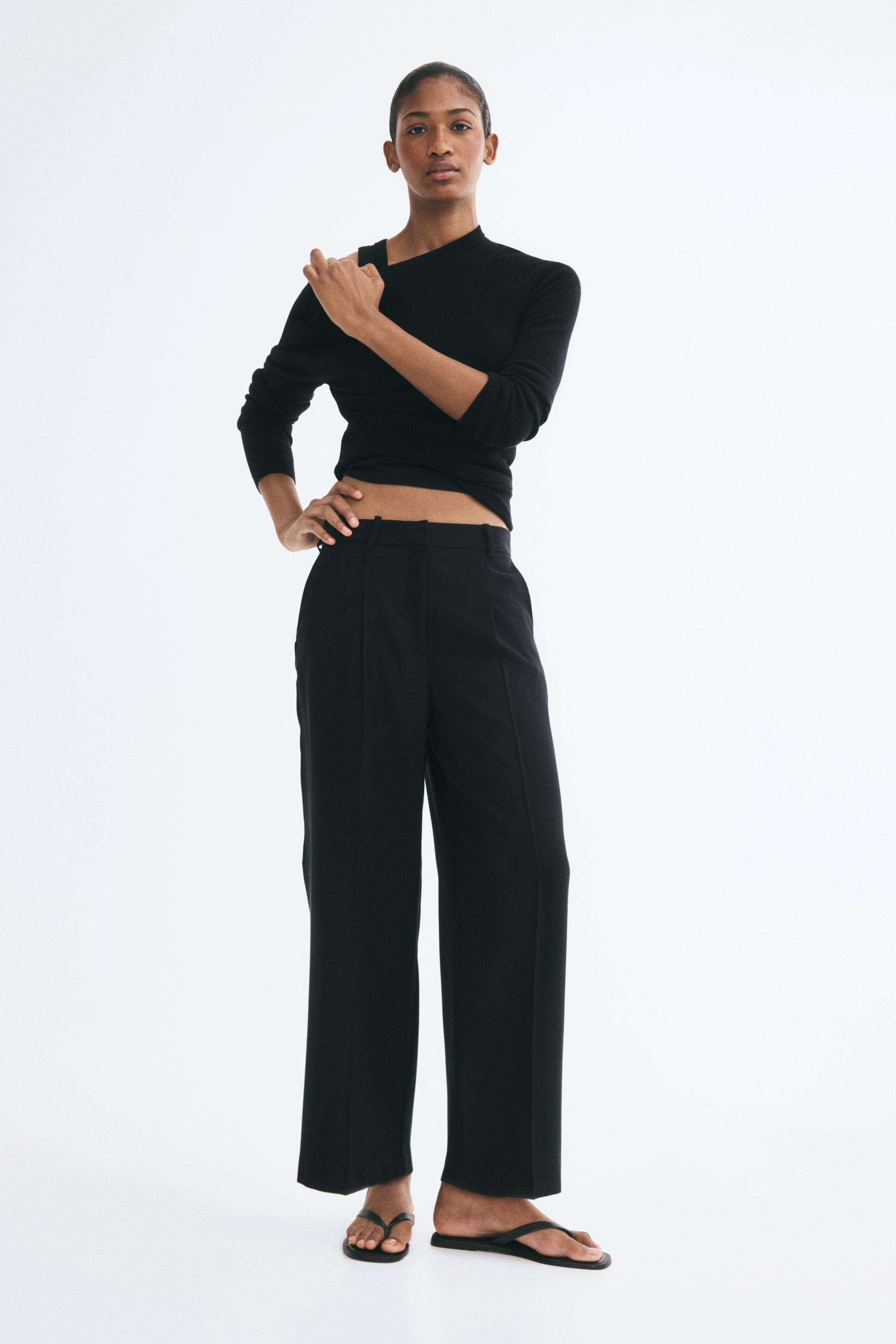 View larger image: The model is wearing black, high-waisted, ankle-length straight-leg pants with pressed front creases, standing with one hand on the hip. The pants are styled with a black long-sleeve top and black flat thong sandals.