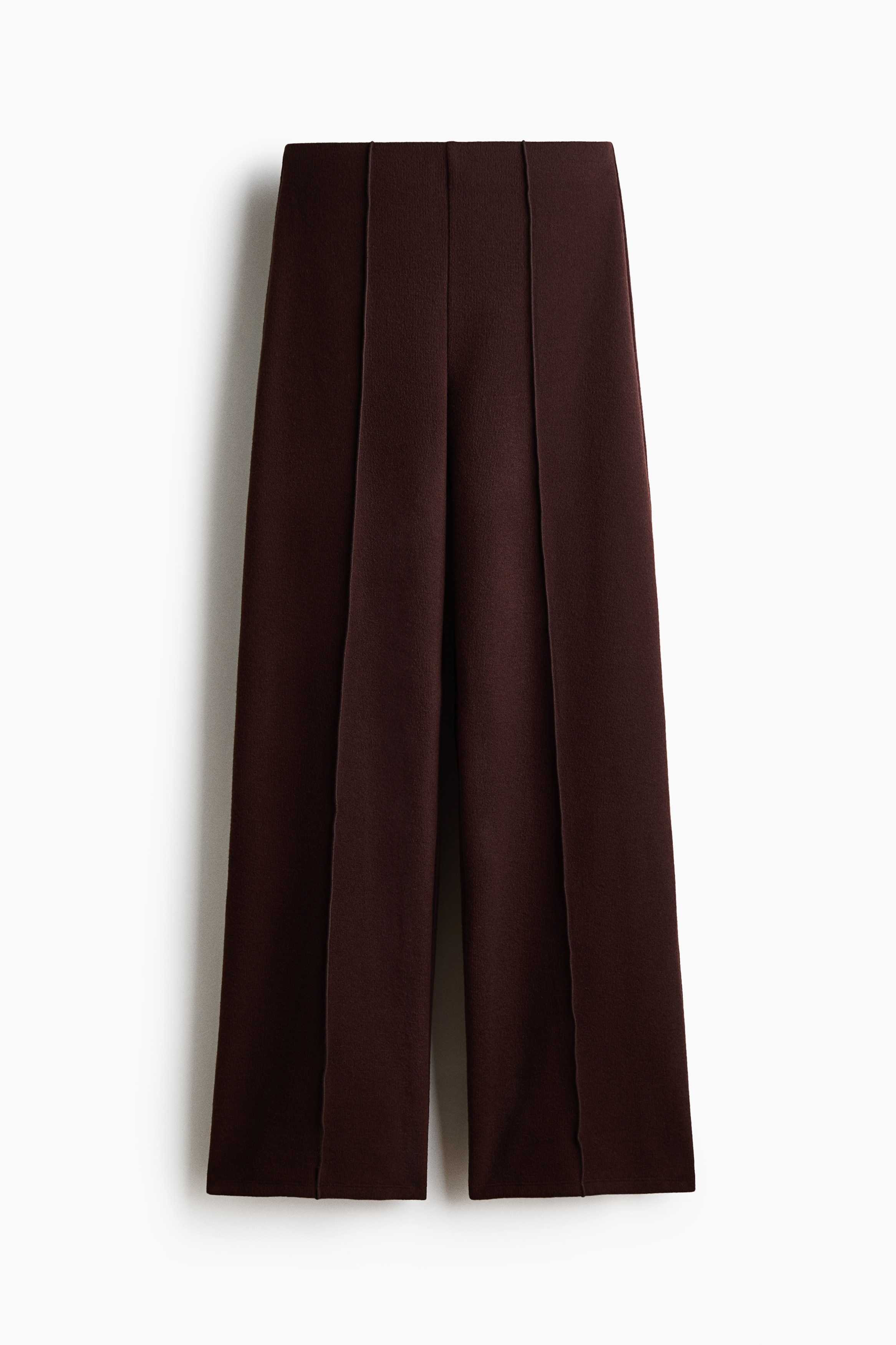 Loose-fit, dark brown knitted trousers featuring straight legs with sewn-in creases down the front are worn by the model, who is sitting on the floor. The trousers are styled with a matching dark brown high-neck top and off-white shoes.