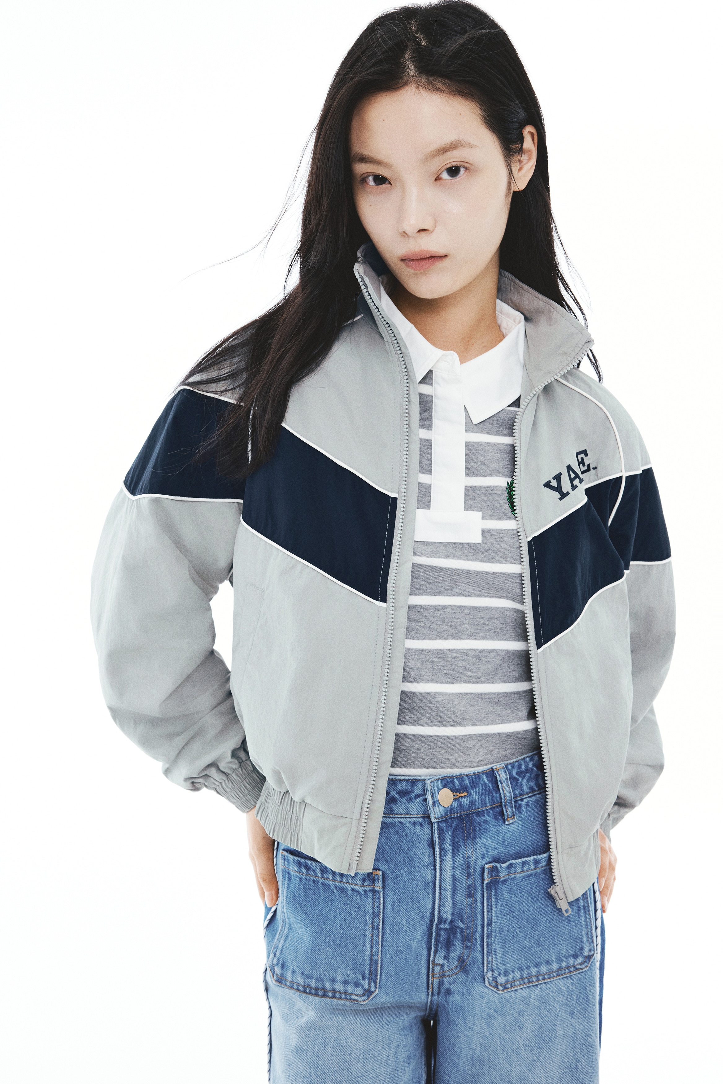 A light gray track jacket is shown from the front, featuring a navy blue chevron-style color-block panel outlined with white piping across the chest and sleeves. The jacket has a stand-up collar, a full-length front zipper, elasticized cuffs and hem, and the word "YALE" embroidered on the left chest.
