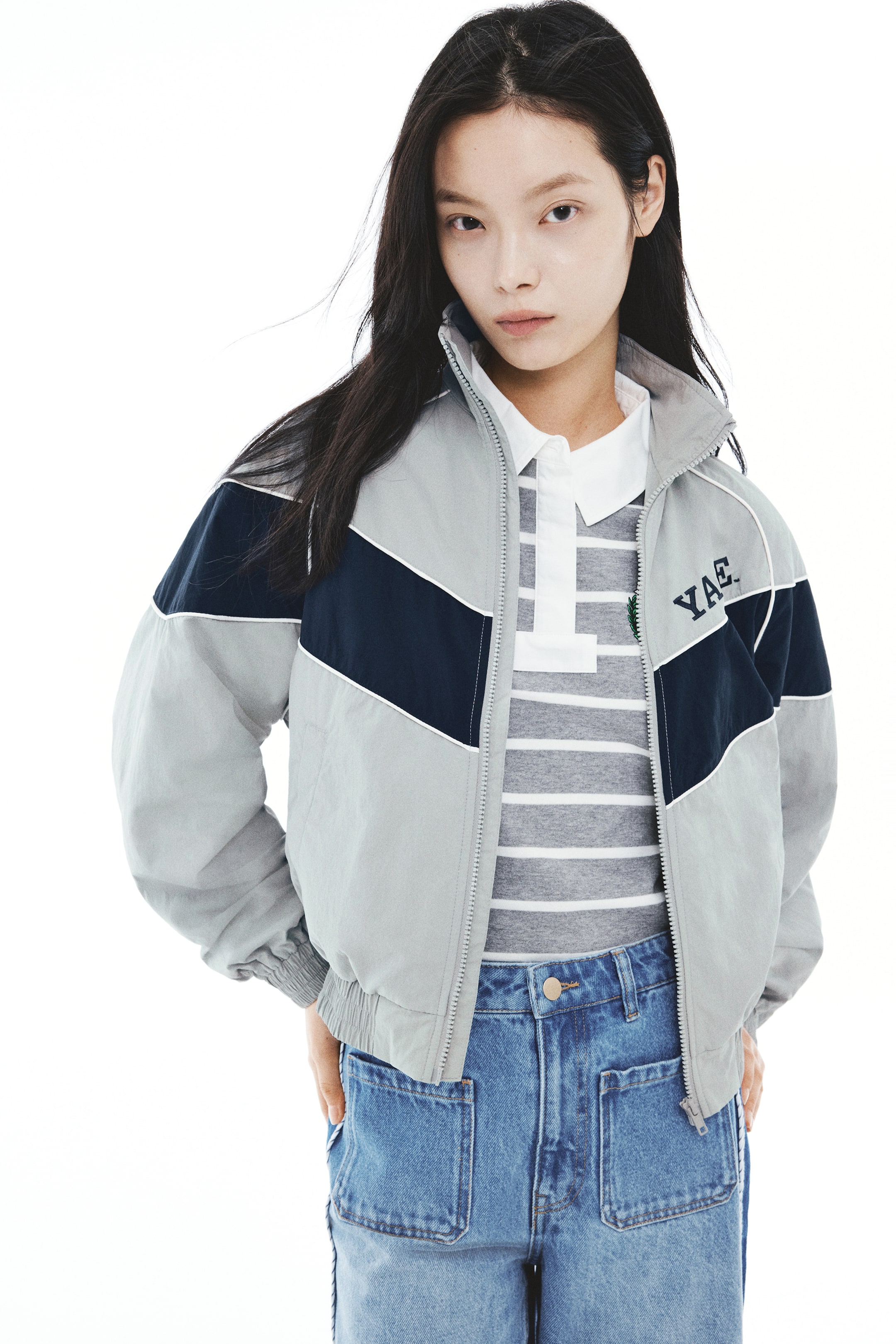 View larger image: A light gray nylon track jacket featuring a navy blue color-blocked chest panel with white decorative piping and embroidered lettering on the chest is being worn open by the model. The jacket, designed with a stand collar, raglan sleeves, and a smocked elastic hem, is layered over a striped polo shirt and paired with blue jeans.