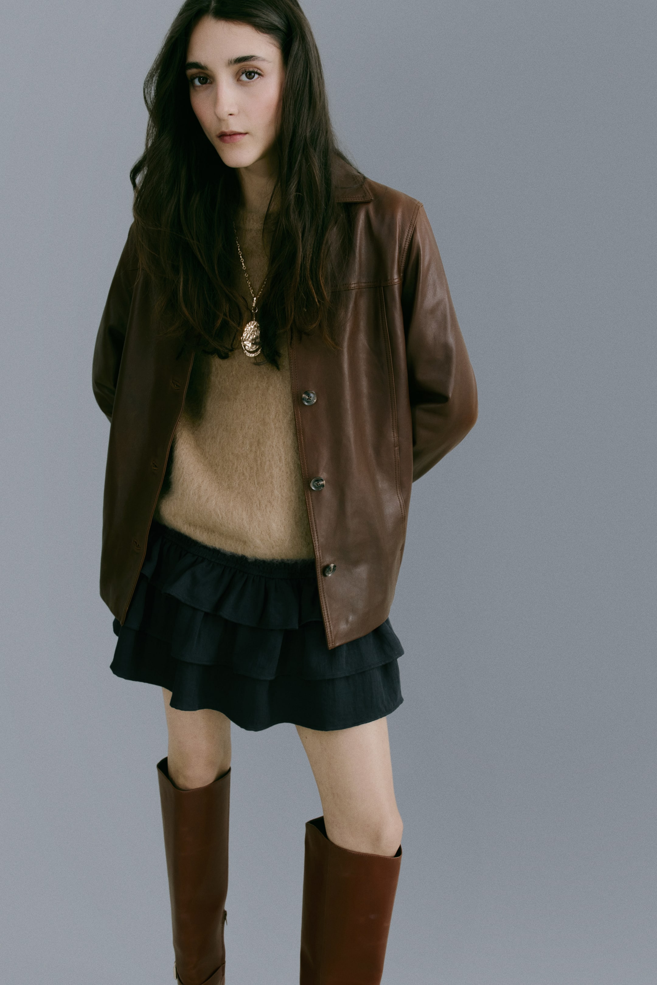 Ver imagen más grande: La modelo lleva un skort de color oscuro, corto y de ajuste holgado, que presenta un amplio fruncido elástico en la cintura y volantes en capas. El skort está combinado con un suéter marrón claro y una chaqueta de cuero marrón.