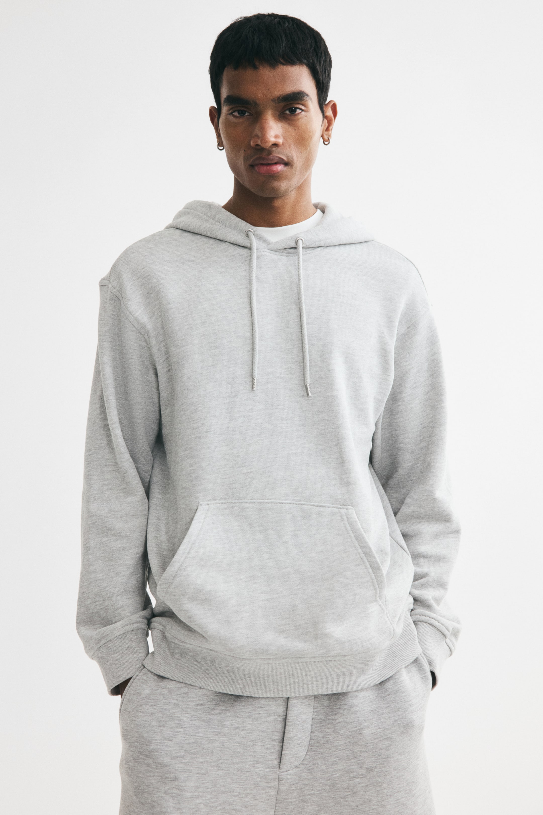 View larger image: Model sedang memakai hoodie kelabu muda dengan potongan santai, bahu jatuh, hud bertali, poket kanggaru, serta manset dan hem berusuk. Tangan model diletakkan dalam poket kanggaru, dan t-shirt putih kelihatan di garis leher, dengan seluar sukan kelabu muda yang sepadan turut dipakai.