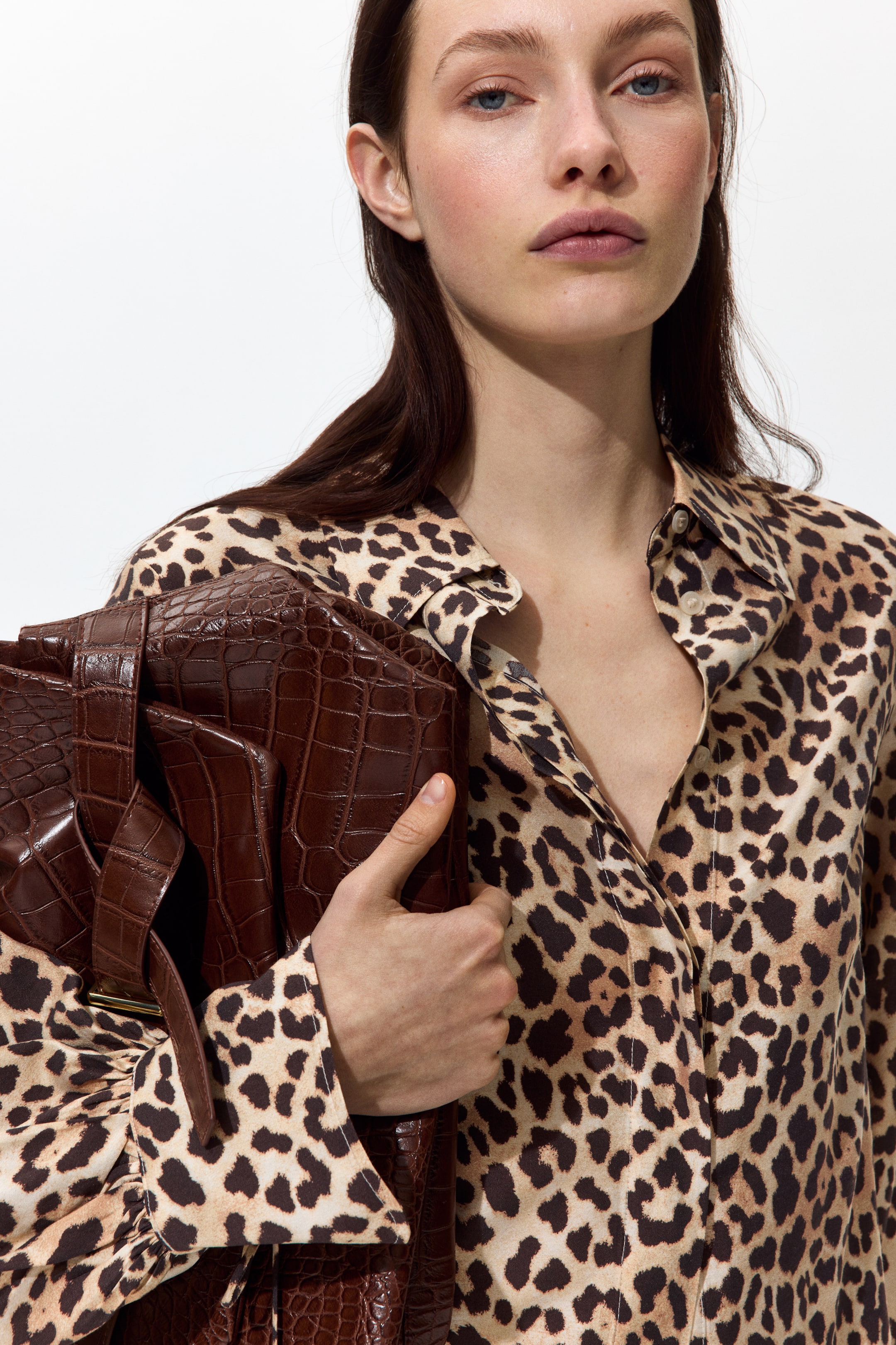 View larger image: A leopard print collared shirt is being worn, featuring a beige base with dark brown spots. Its front placket is partially unbuttoned, and a long sleeve with a buttoned cuff is gently creasing from being held.
