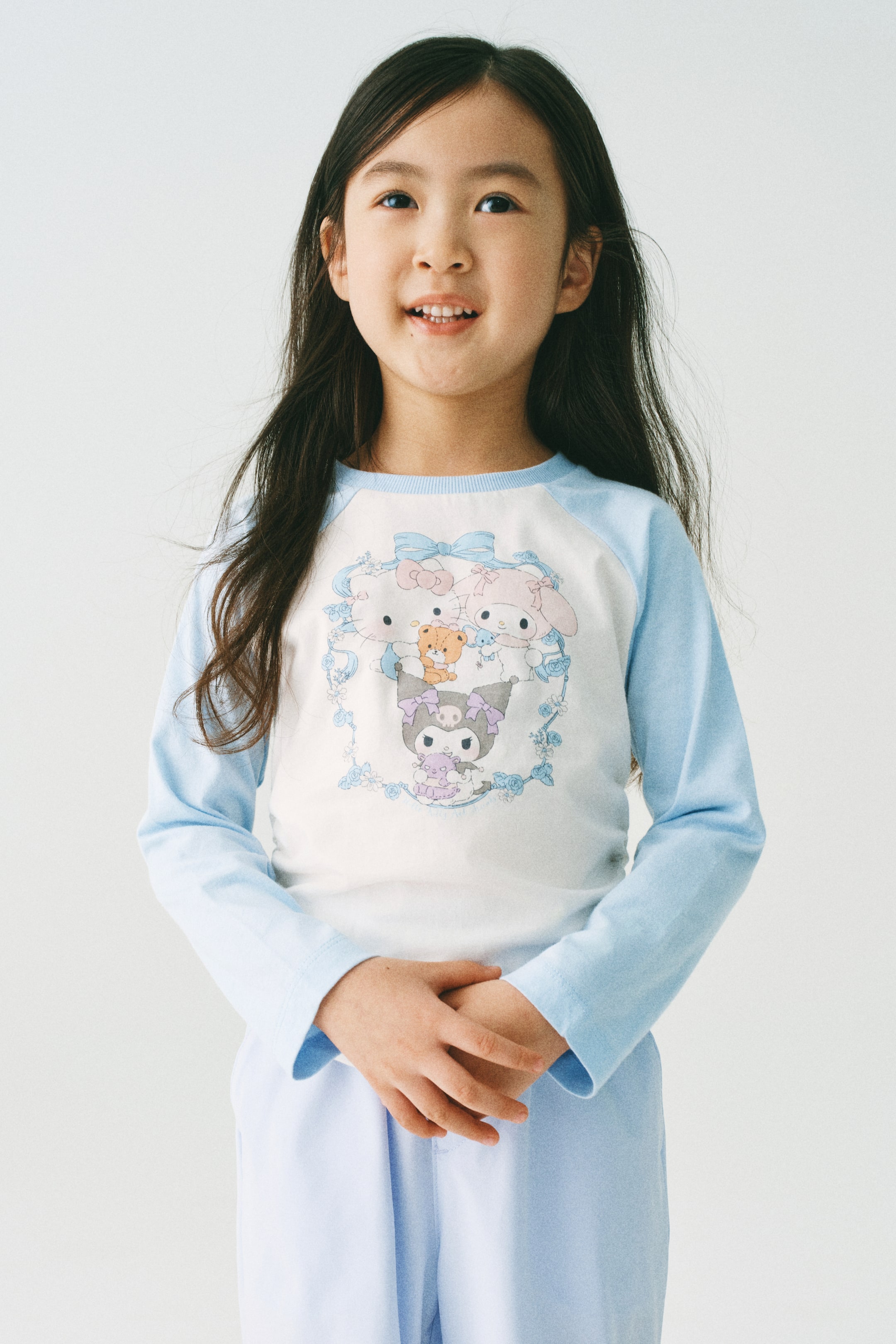 이미지 확대 보기: 모델이 착용한 프린트된 면 저지 탑은 둥근 리브 트리밍 넥라인과 긴 연한 파란색 래글런 소매, 그리고 흰색 앞면에 다채로운 만화 캐릭터 프린트를 특징으로 합니다. 모델의 손은 앞쪽에 함께 놓여 있습니다.