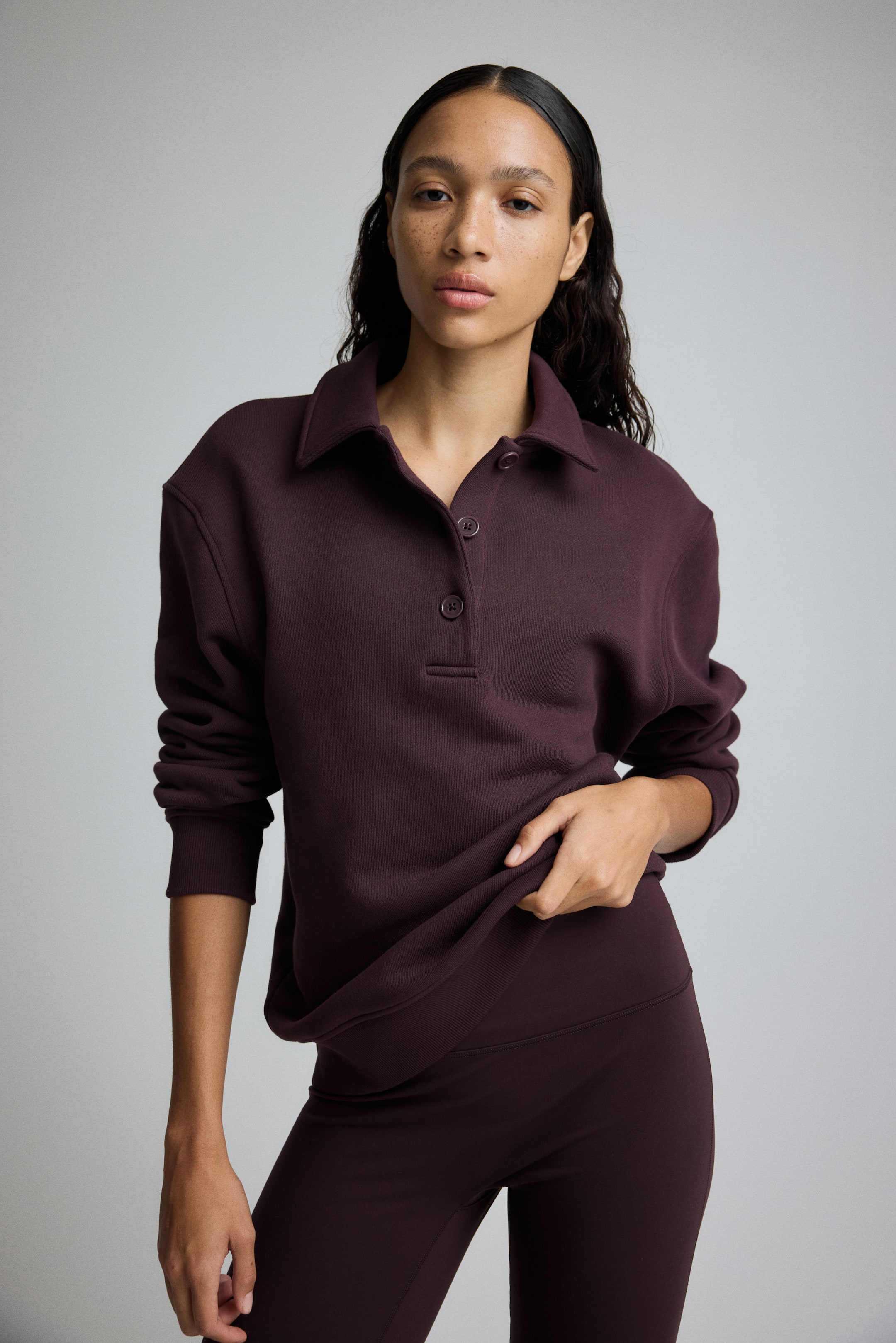 Grotere afbeelding bekijken: Een donker kastanjebruine polo sweatshirt wordt van voren gepresenteerd, met een klassieke kraag, een drieknoopssluiting en lange mouwen met geribbelde manchetten. De geribbelde zoom wordt omhoog gehouden, waardoor bijpassende donker kastanjebruine leggings te zien zijn die ook worden gedragen.