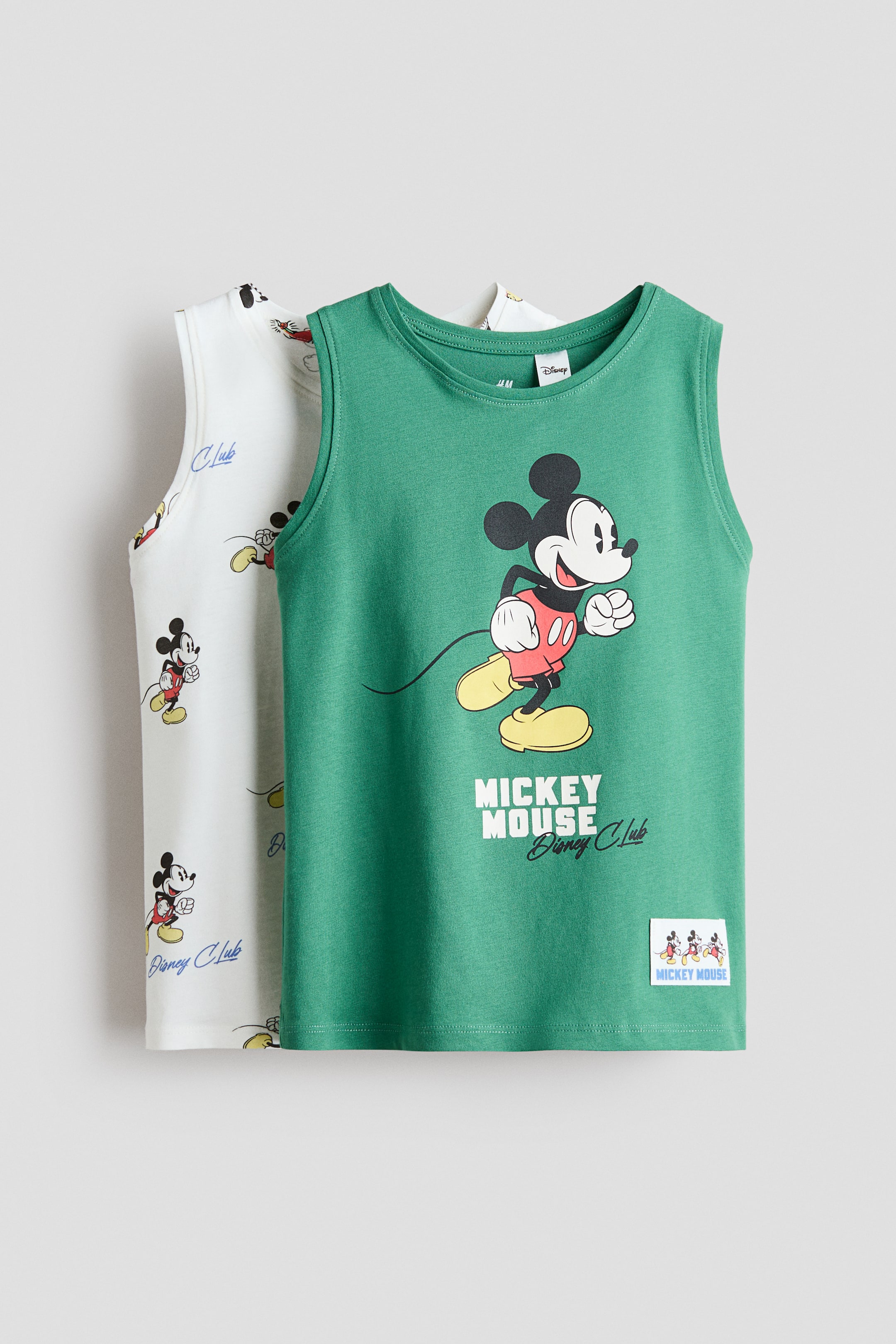Visualizza l'immagine ingrandita: Due canottiere sono esposte: la canottiera verde davanti presenta una grande grafica di Topolino in posa da corsa, accompagnata dal testo "Mickey Mouse Disney Club" e una piccola etichetta rettangolare sull'orlo. La canottiera bianco sporco parzialmente visibile dietro mostra un motivo ripetuto di figure più piccole di Topolino e il testo "Disney Club".