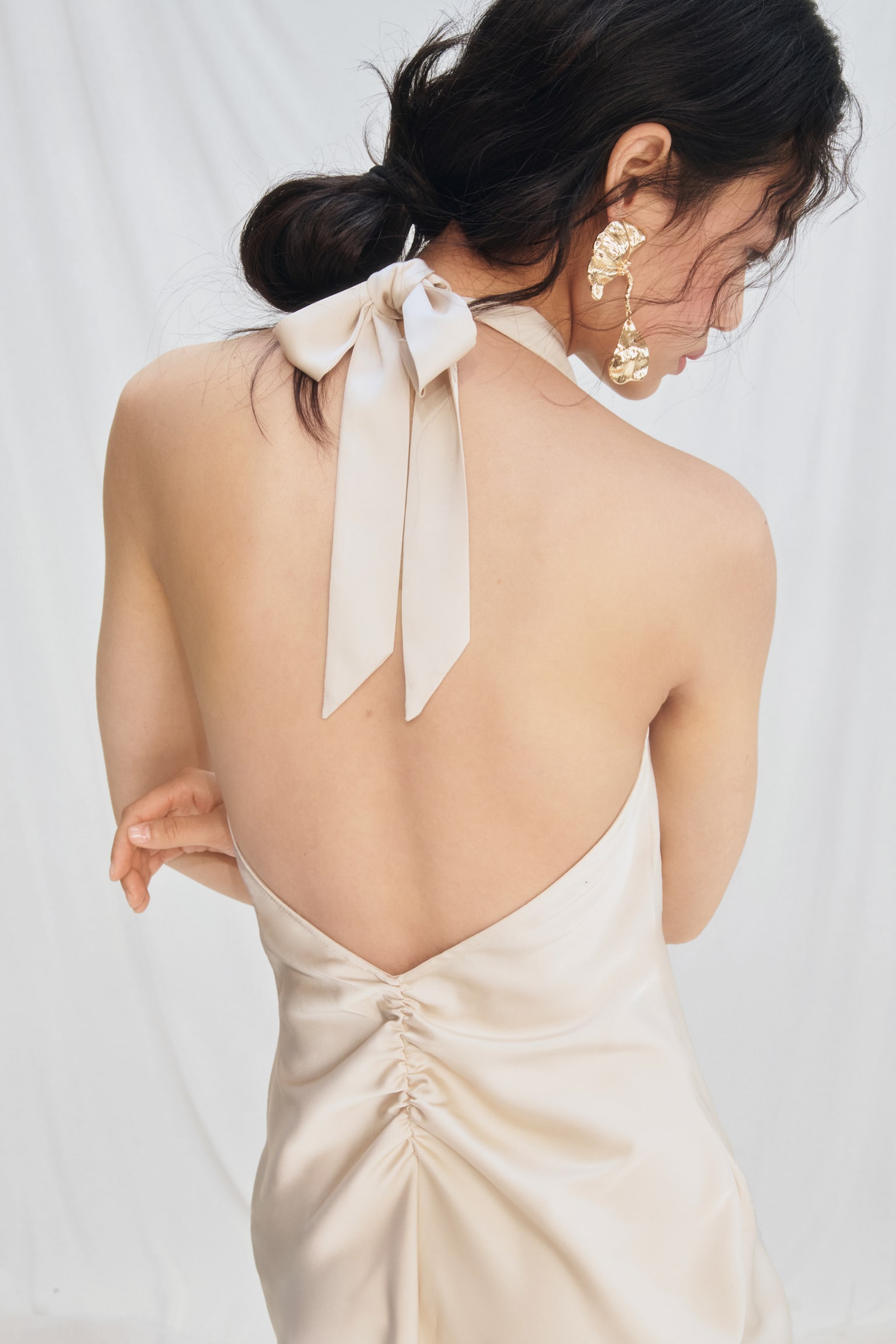 The model is wearing a softly draping satin dress, featuring wide halterneck straps that tie into a bow at the back of the neck. The dress boasts an open back with a gathered seam at the lower back, which the model's hand is gently touching. - 1