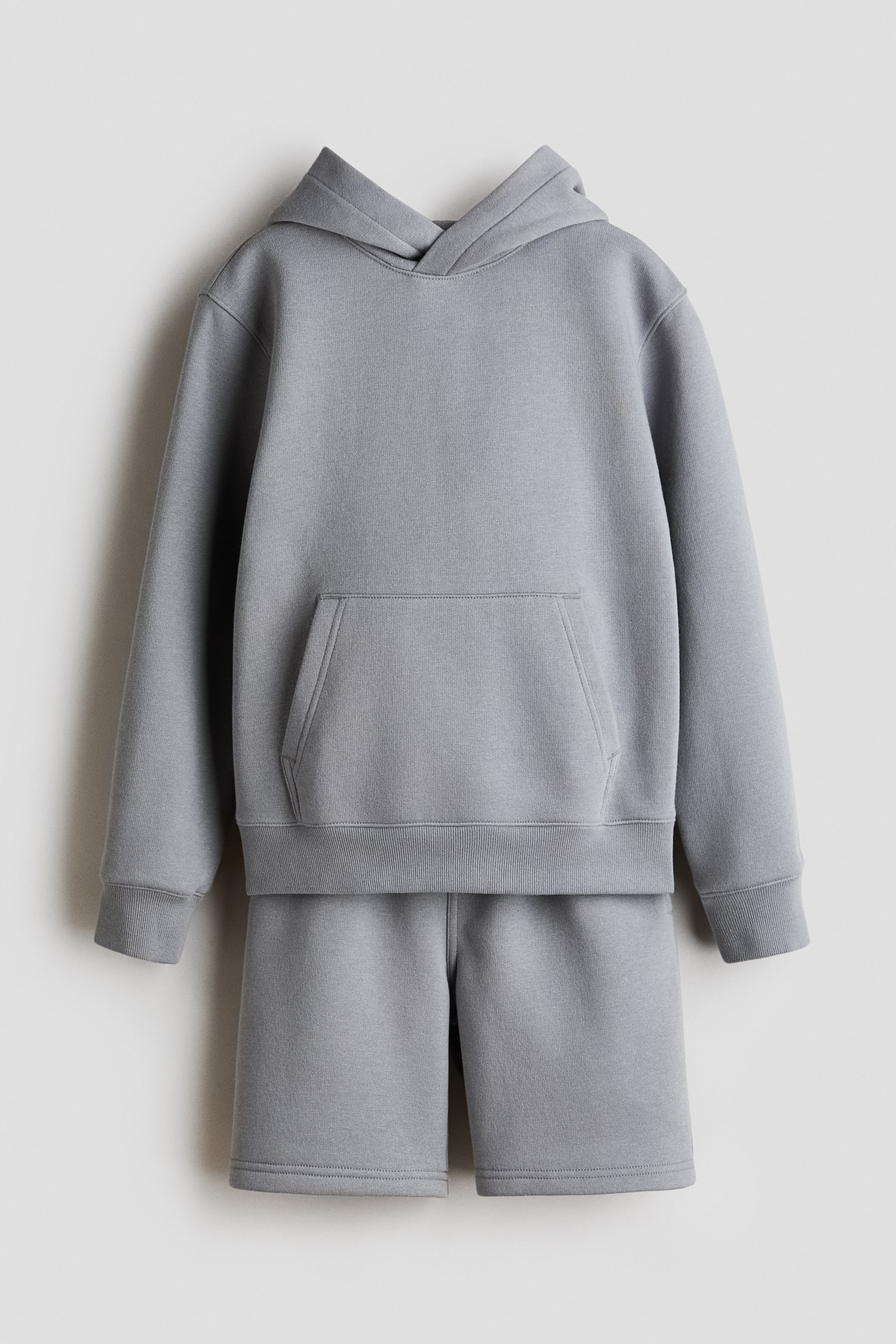Visualizza immagine più grande: Un set composto da felpa grigia con cappuccio e pantaloncini coordinati è presentato nella sua vista frontale. La felpa presenta un cappuccio, maniche lunghe con polsini a coste, una tasca a marsupio e un orlo a coste, mentre i pantaloncini mostrano un tessuto grigio coordinato con orli delle gambe rifiniti.