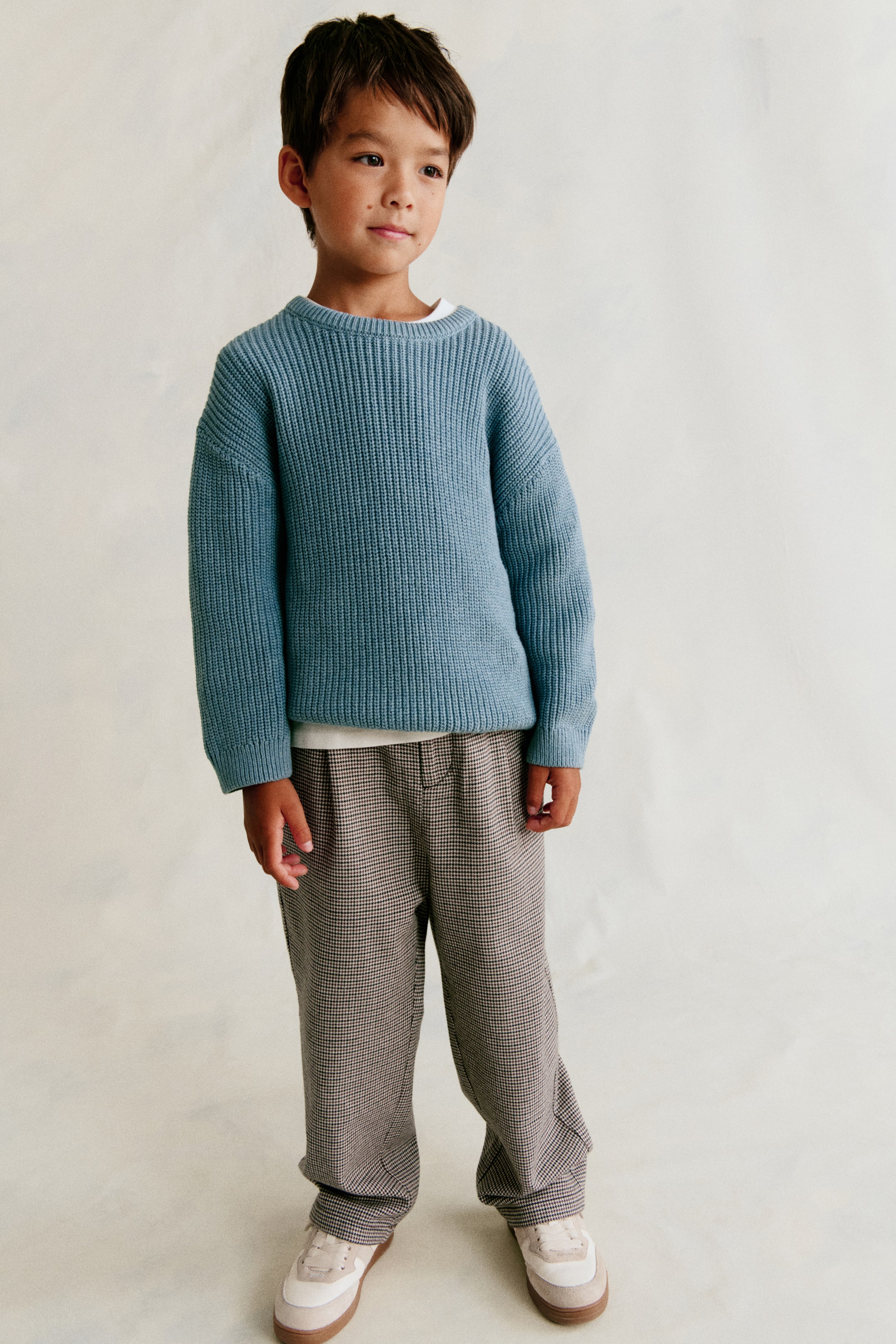 View larger image: The chinos feature a brown and off-white small check pattern, with an elasticated waist and visible button closure, plus front pleats. They are shown in a relaxed straight-leg fit with diagonal side pockets.