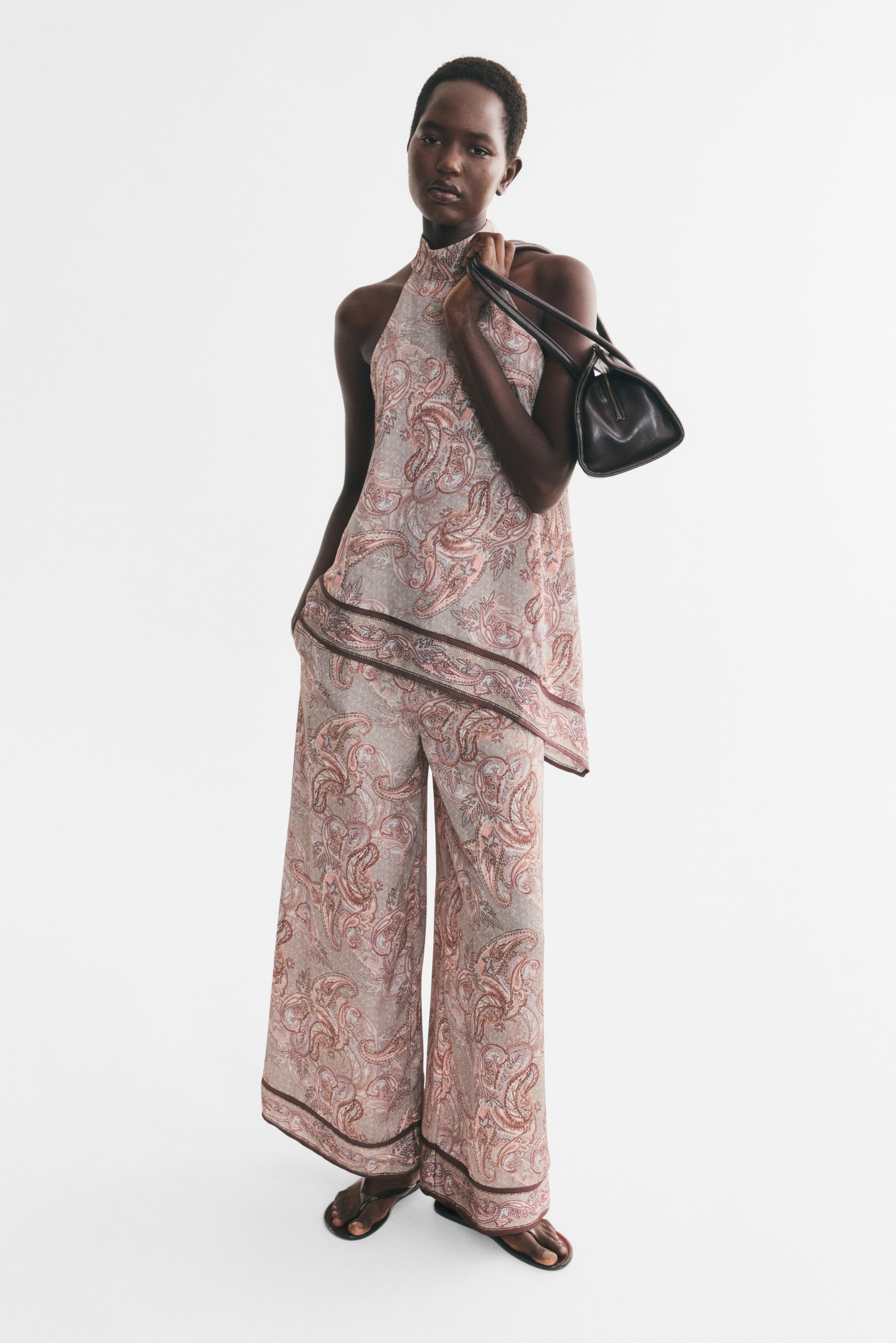 View larger image: Wide-leg pants in a woven fabric with a pink paisley pattern and a slight sheen are being worn by the model. The pants feature a dark border at the hem and are styled with a matching halter-neck top, with the model placing one hand in a side pocket while carrying a dark brown shoulder bag.