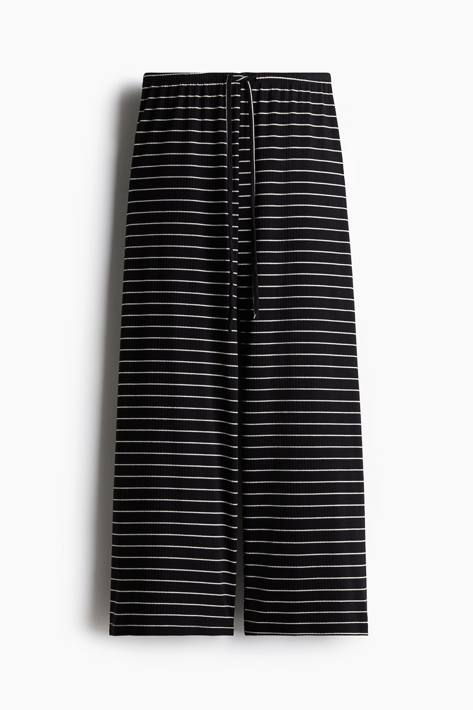 Le pantalon présente un tissu noir avec de fines rayures horizontales blanches et une texture côtelée. Il est doté d'une taille élastique avec un cordon de serrage blanc et arbore une coupe large.