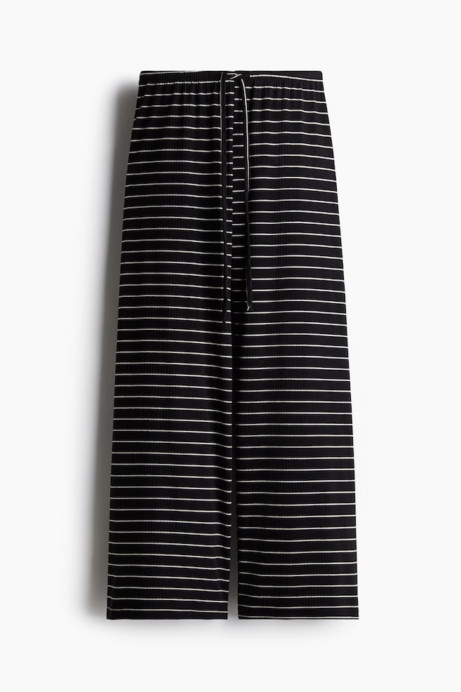 Los pantalones muestran una tela negra con finas rayas horizontales blancas y una textura acanalada. Presentan una cintura elástica con un cordón blanco y un estilo de pierna ancha. - 1
