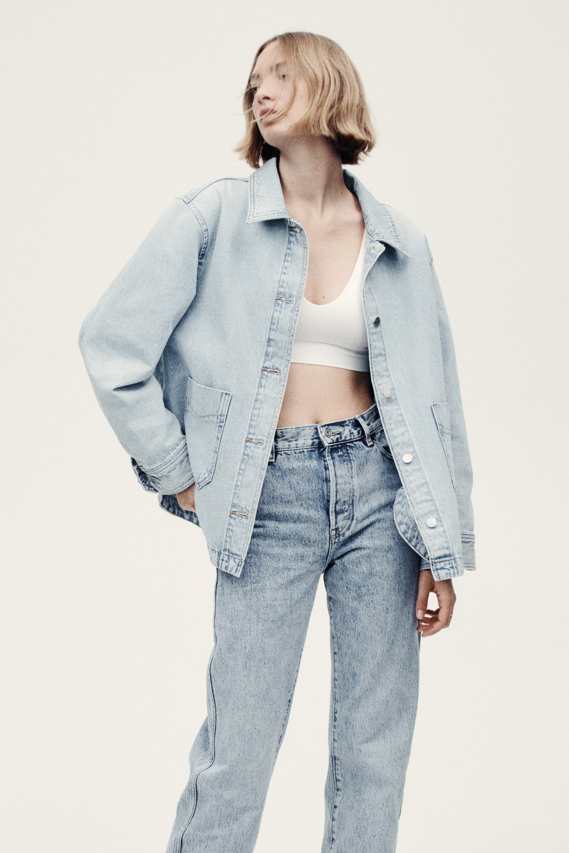 View larger image: The model is wearing a light blue rigid denim shacket, featuring a collar, an open front with buttons, long sleeves with buttoned cuffs, and patch front pockets. She is placing her right hand into one of the front pockets, while a white bra top is visible underneath, complemented by light wash denim jeans to complete the look.