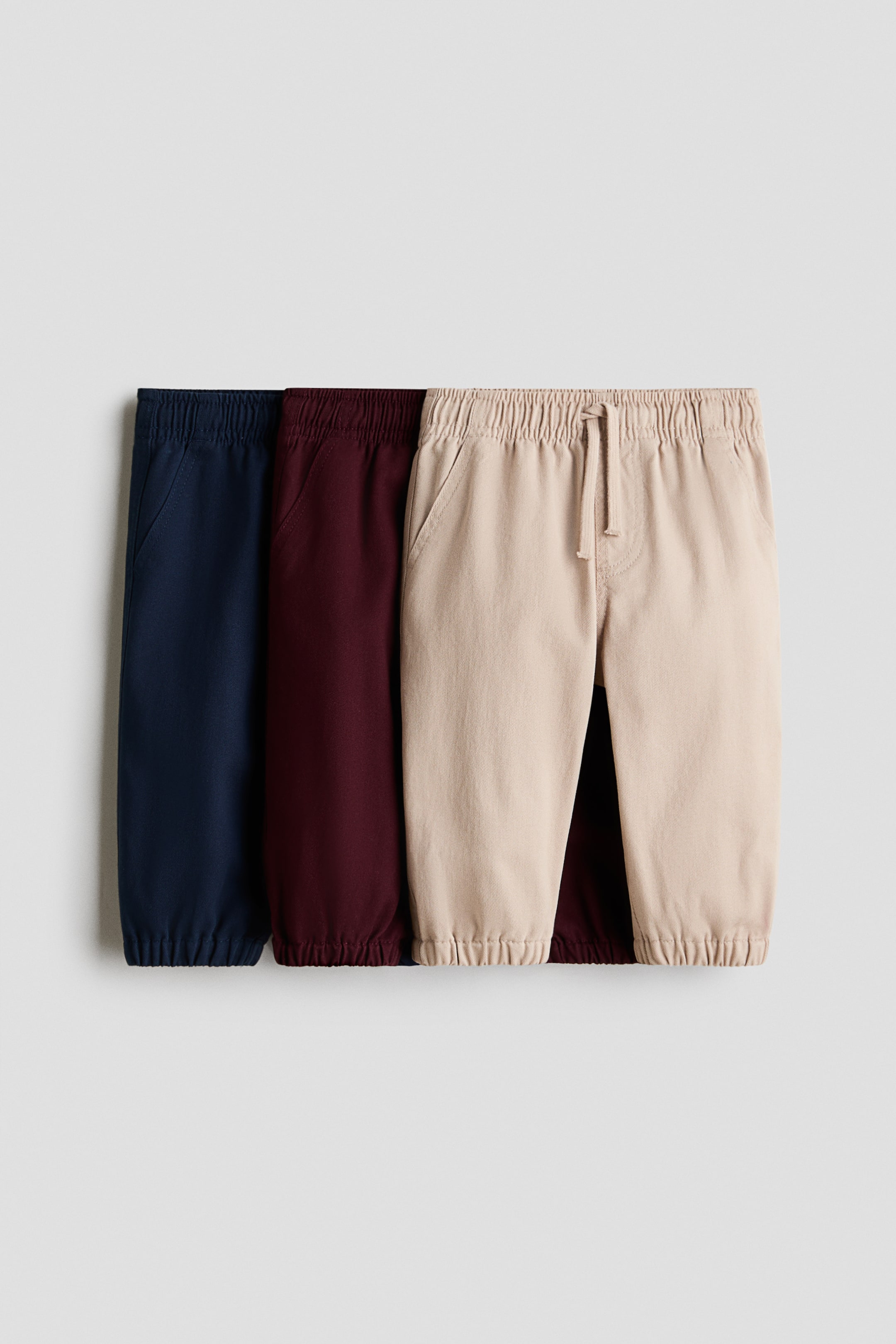 View larger image: Three pairs of jogger pants in beige, maroon, and navy blue are laid out flat, overlapping from front to back, showcasing elasticated waistbands and cuffs. The front beige pair features a drawstring and front pockets.