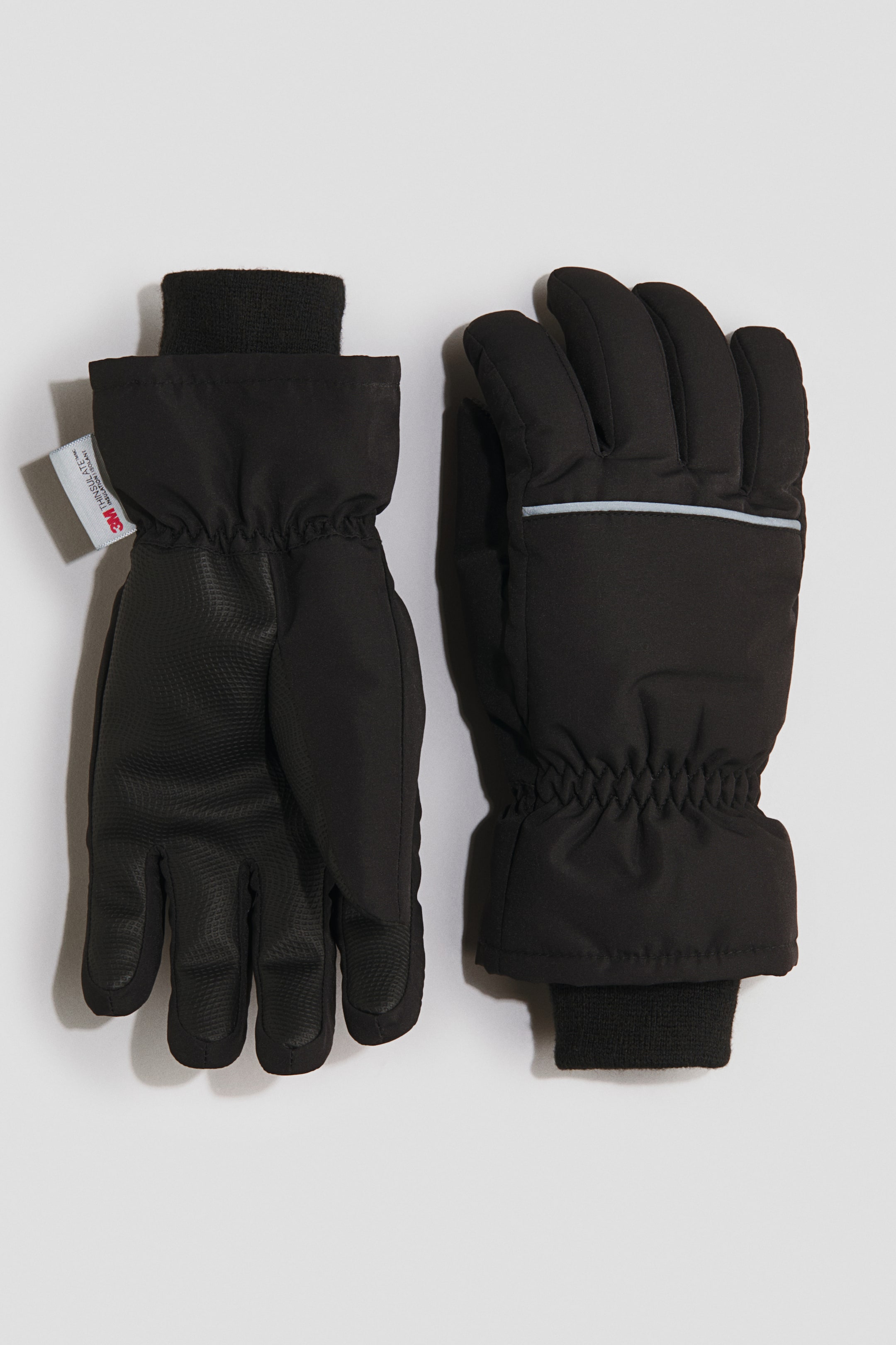 Grösseres Bild ansehen: Schwarze Handschuhe liegen flach, wobei einer die strukturierte Handfläche und der andere den gepolsterten Handrücken zeigt; beide verfügen über elastische Handgelenke und schwarze gerippte Strickbündchen. Der linke Handschuh zeigt ein weisses Etikett mit «3M Thinsulate», und der rechte Handschuh zeigt einen hellblauen horizontalen Streifen über den Handrücken.