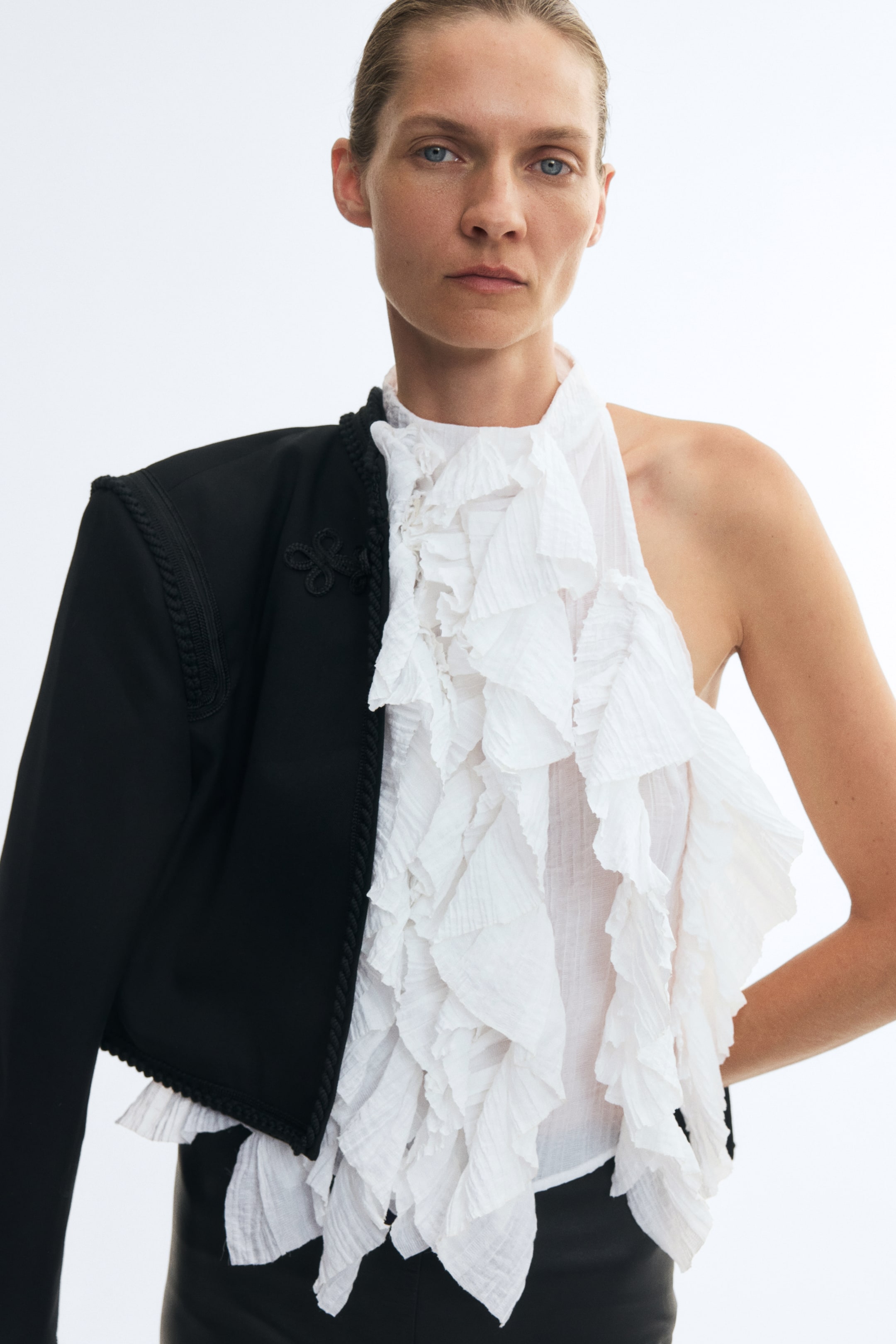 View larger image: The model is wearing a white sleeveless top featuring cascading pleated ruffles with raw edges and a high round neckline. A cropped black jacket with braided trim is draped over the model’s right shoulder, leaving the other side exposed.