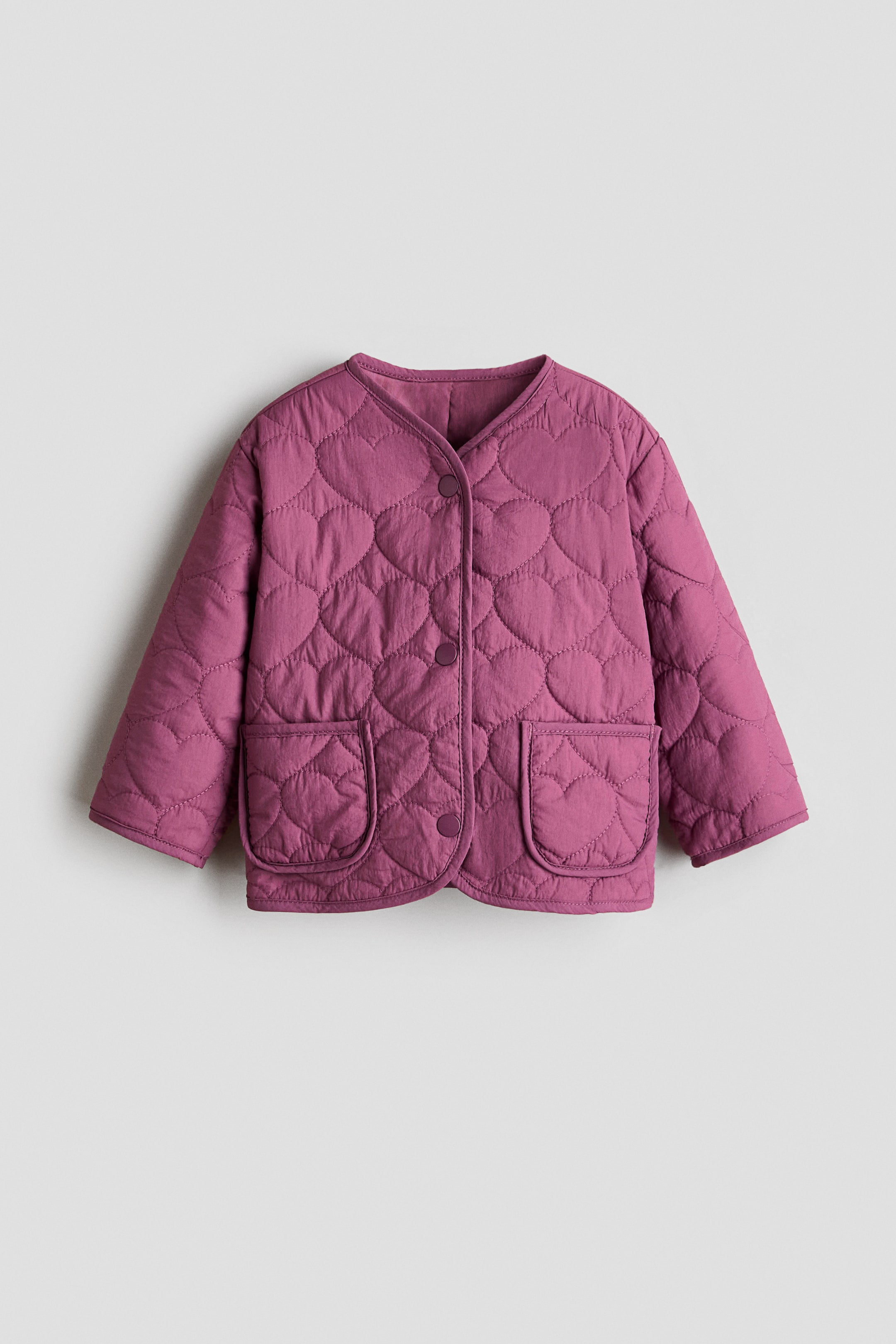 View larger image: A mauve quilted jacket showcasing a heart-shaped quilting pattern throughout, complemented by a V-neckline and a front button closure with four matching buttons. It also features two rounded patch pockets on the front and is displayed facing forward.