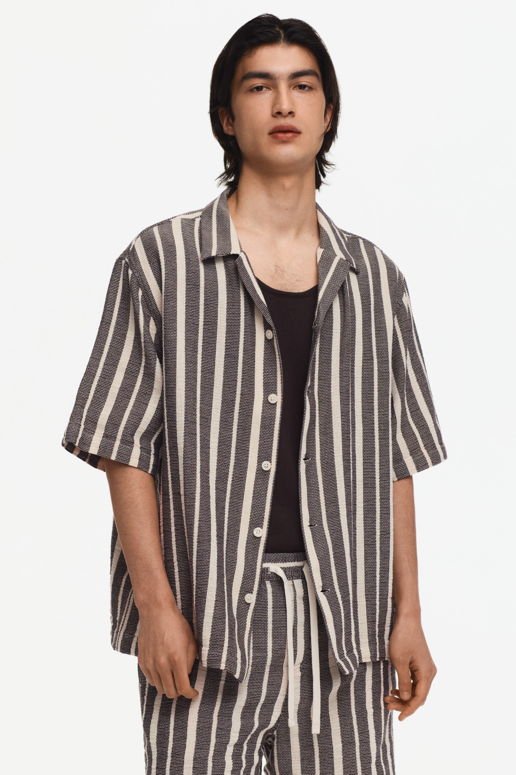 הצגת תמונה גדולה יותר: חולצה עם צווארון קמפ ומכנסיים קצרים תואמים עם שרוולים קצרים, מעוטרים בפסים אנכיים בגווני אפור כהה ולבן שבור עם אריגת טקסטורה. החולצה מוצגת מלפנים, פתוחה, וחושפת חולצת בסיס שחורה, בעוד המכנסיים מציגים מותן עם שרוך לבן.