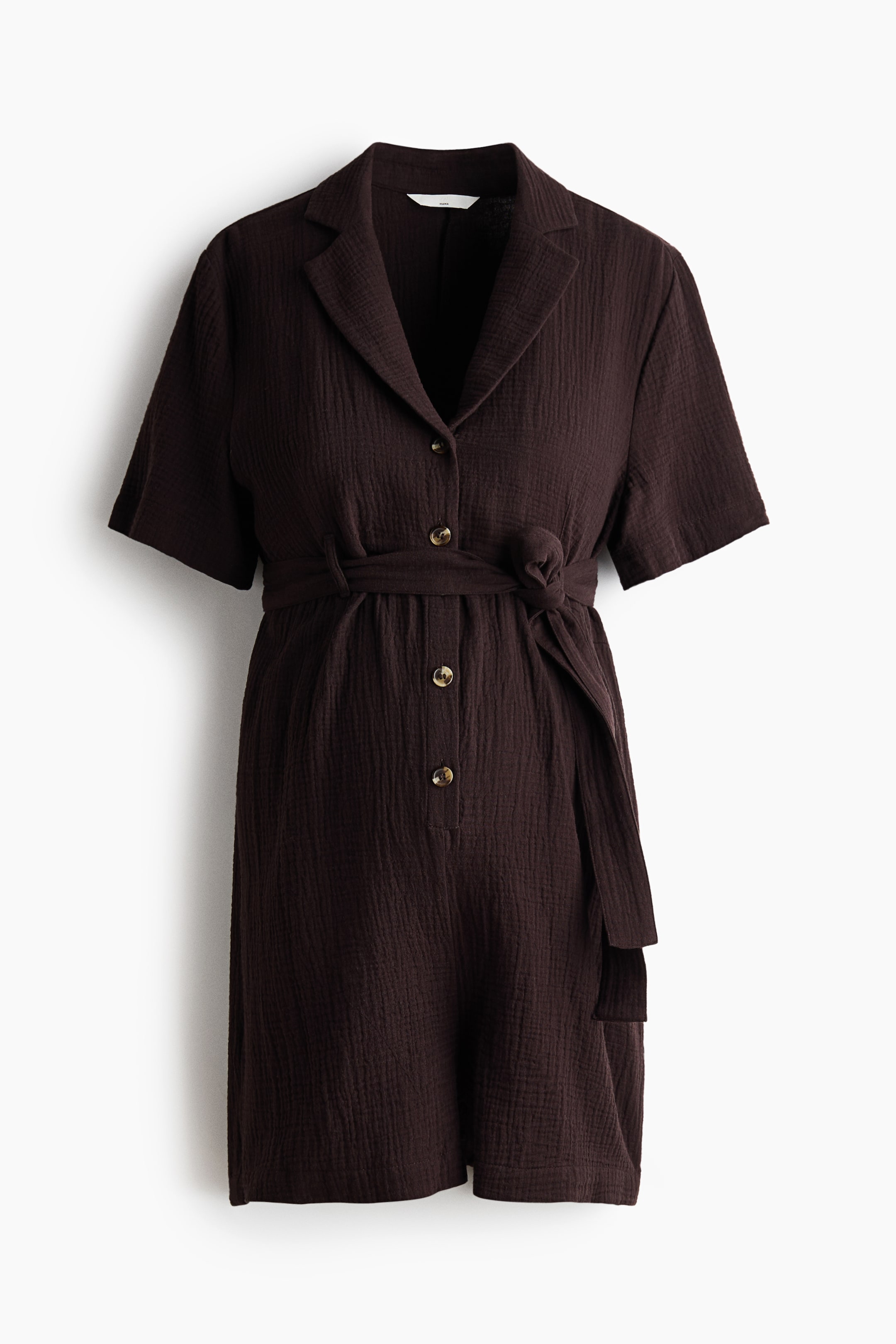 View larger image: The front view showcases a dark brown short-sleeved jumpsuit, highlighting a collared neckline with notch lapels and a full-length button placket adorned with light brown buttons. A self-fabric tie belt cinches the waist, knotted at the front, all crafted from a textured, crinkled fabric.