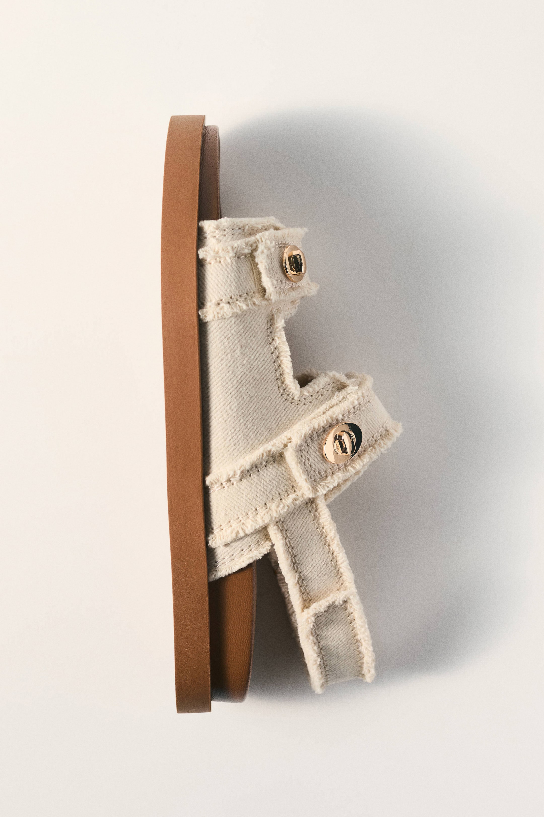 Vezi imaginea mai mare: Un sandal de culoare crem este poziționat pe lateral, afișând un material textil împletit cu margini destrămate și două încuietori cu mecanism de răsucire de culoare aurie. Este proiectat cu o talpă groasă de culoare maro.