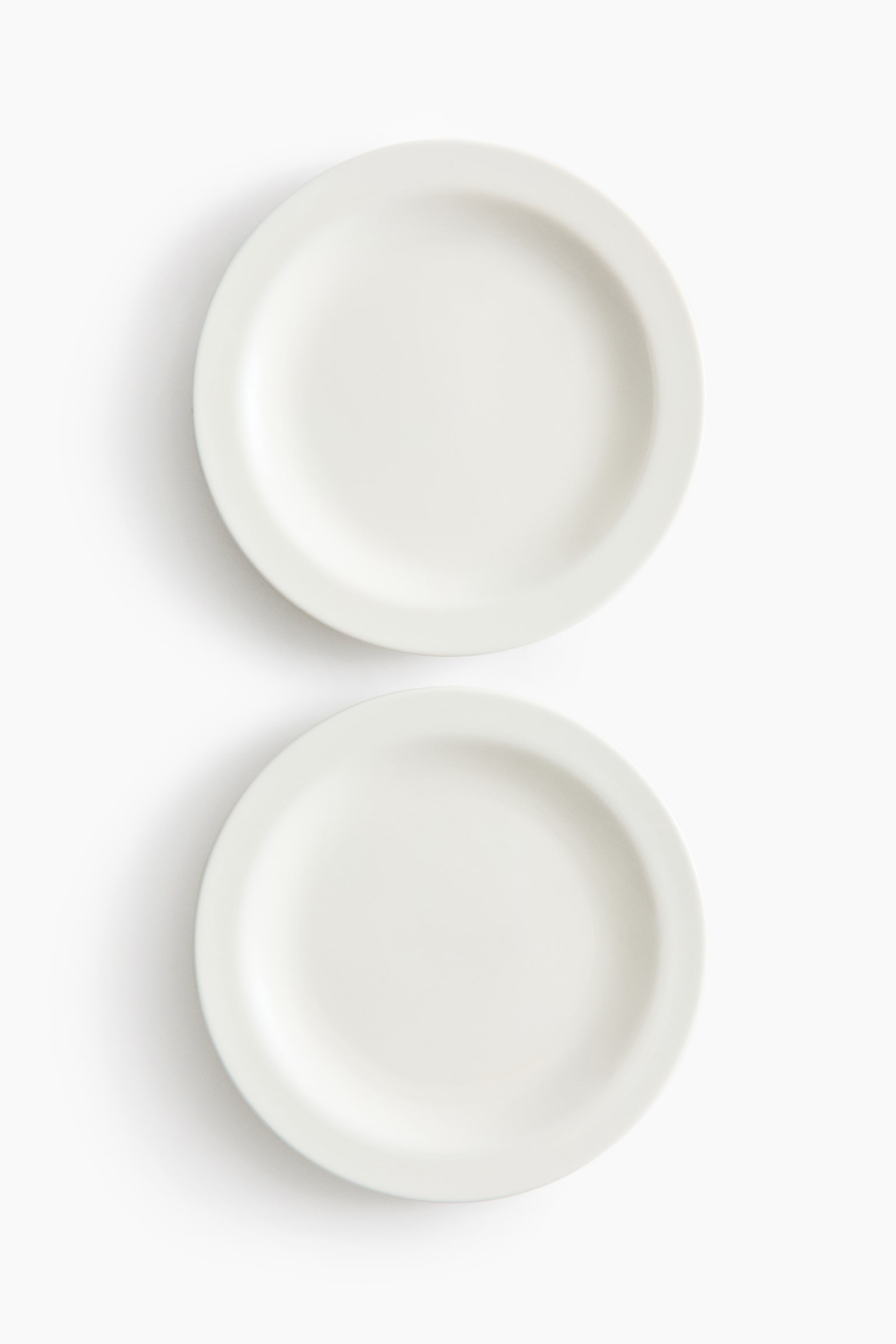 Two round, off-white plates are displayed, each showcasing a wide, subtly curved rim and a recessed central dining surface. They are arranged vertically, with one plate positioned above the other. - 1