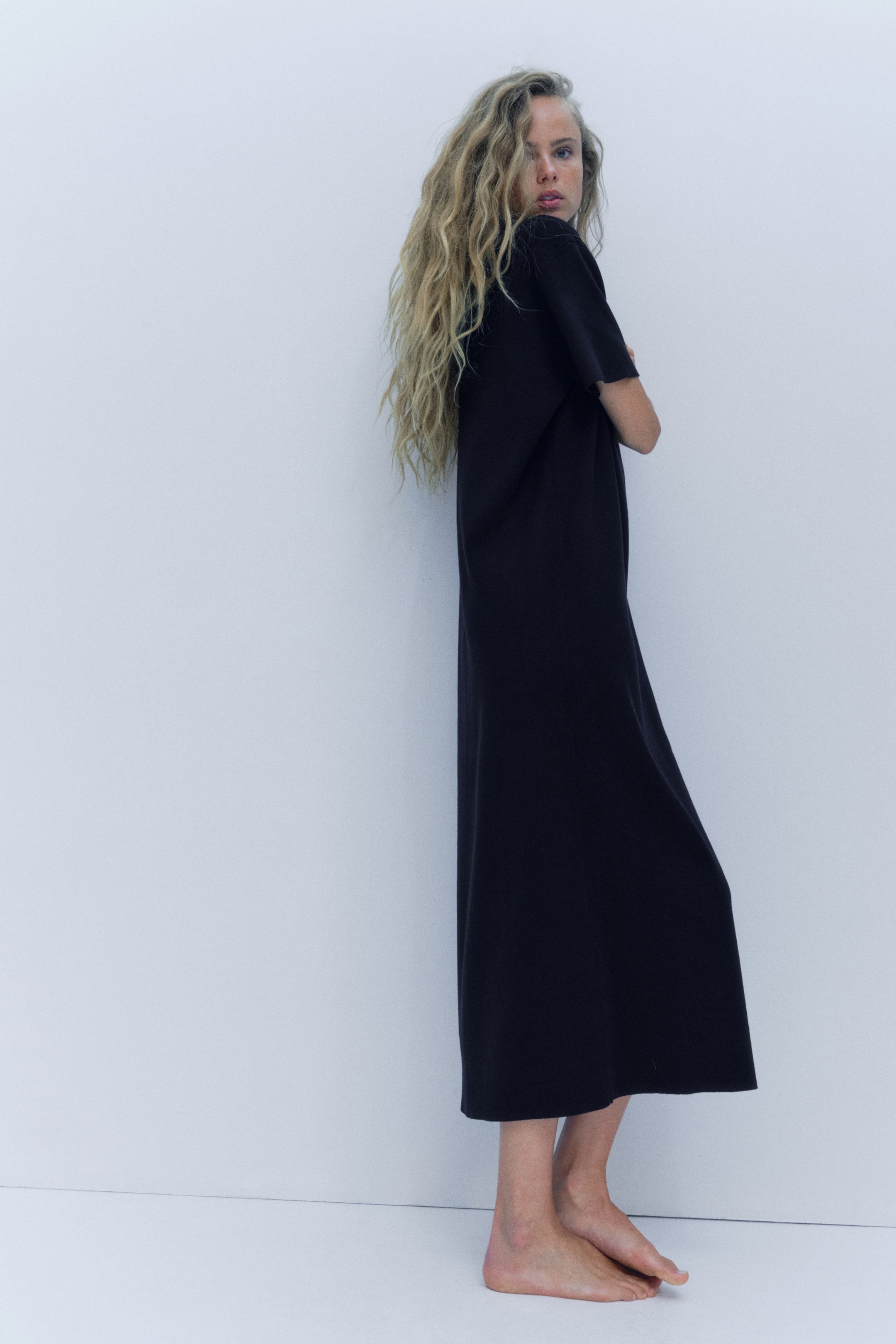 View larger image: The model is wearing an ankle-length oversized black fine-knit dress, shown in a three-quarter back view. The dress features dropped shoulders, short sleeves, and a straight drape that falls to the lower calf.