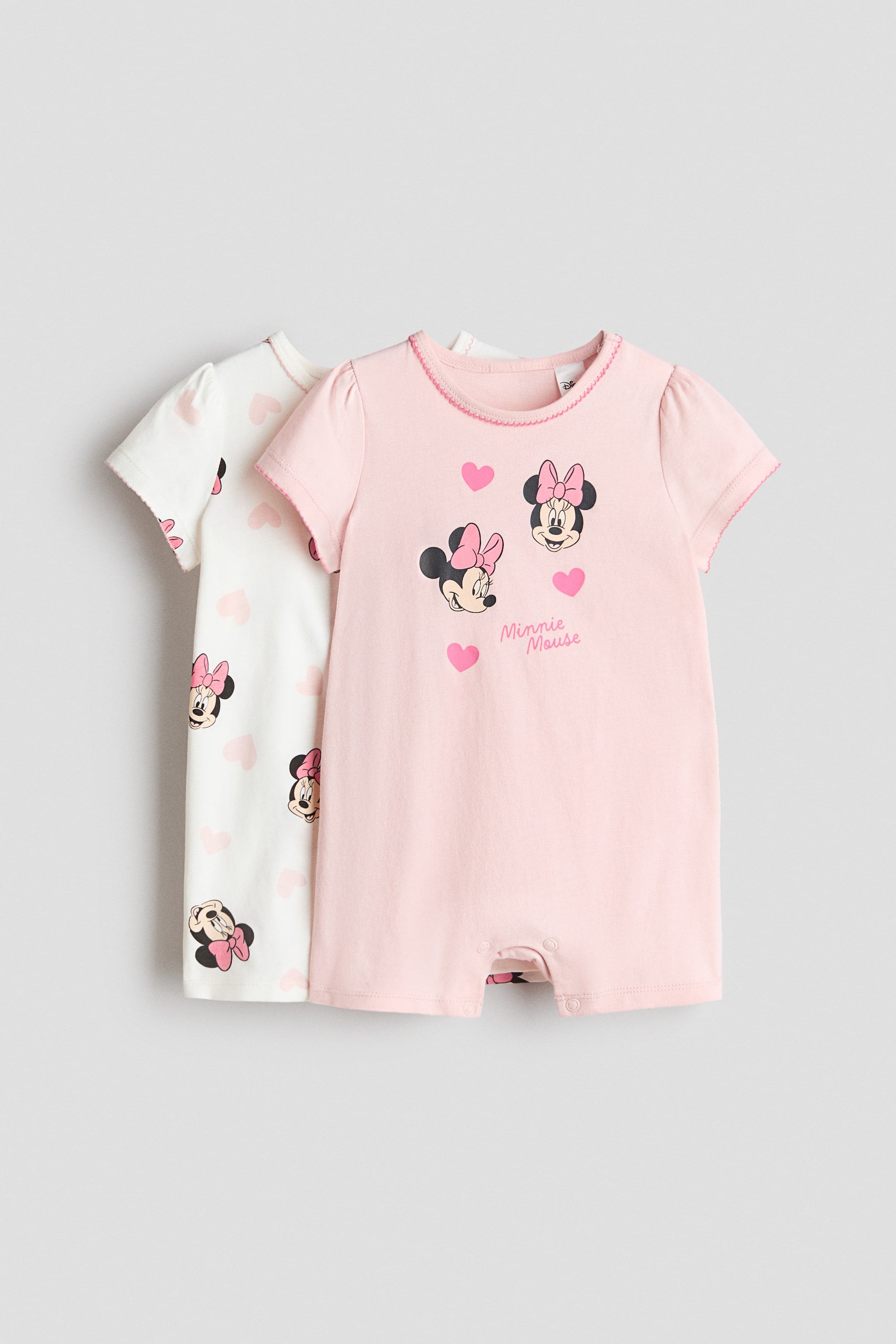 Agrandir l'image: Deux pyjamas à manches courtes présentent leurs vues de face, avec des motifs différents en jersey de coton imprimé doux. Le pyjama rose clair met en avant des visages de Minnie Mouse, des cœurs roses et du texte, tandis que le pyjama blanc affiche un motif intégral de visages de Minnie Mouse et de cœurs roses ; tous deux sont conçus avec des encolures rondes, des manches courtes bouffantes, des jambes courtes, des fermetures à pression et des finitions délicates en picot sur les encolures et les poignets des manches.