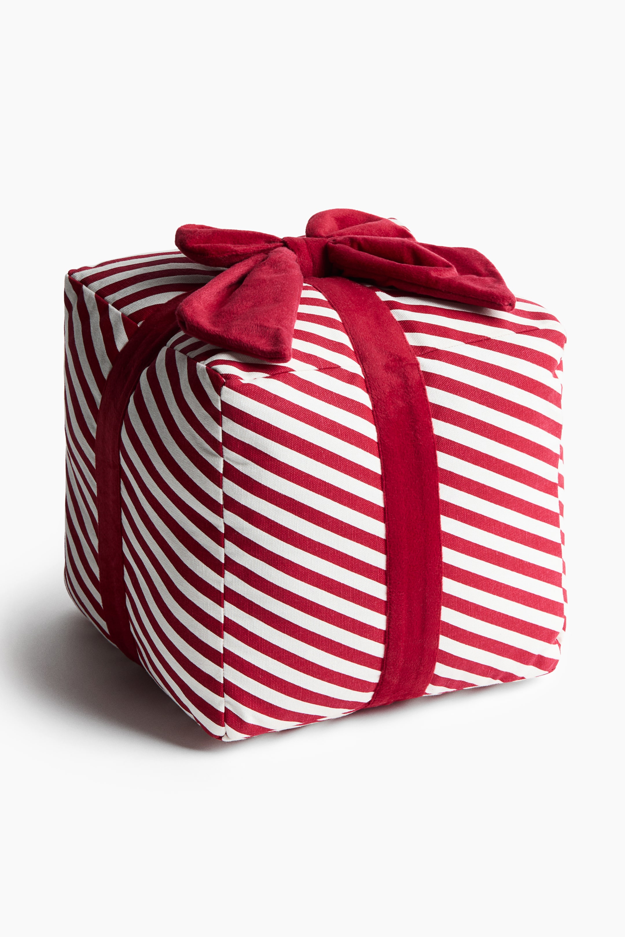 View larger image: A cube-shaped decorative cushion designed to resemble a gift box is shown facing forward at an angle, featuring a red and white diagonally striped pattern. The cushion is wrapped with a thick, plush red ribbon that is tied into a large bow at the top.