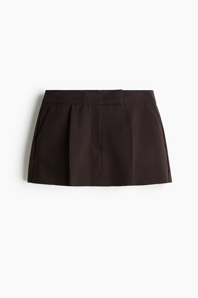 A dark brown mini skort in woven fabric is displayed from the FRONT, featuring an extended waistband, diagonal side pockets, and four pleats at the front. - 1
