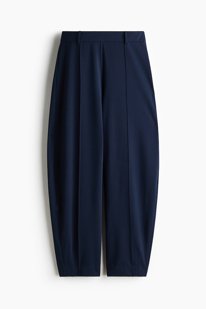 Pantalon bleu foncé présenté en vue de face, avec une ceinture plate dotée de passants et des plis avant cousus. Le pantalon présente une coupe à jambes baril, se rétrécissant vers les chevilles avec un ourlet net. - 1