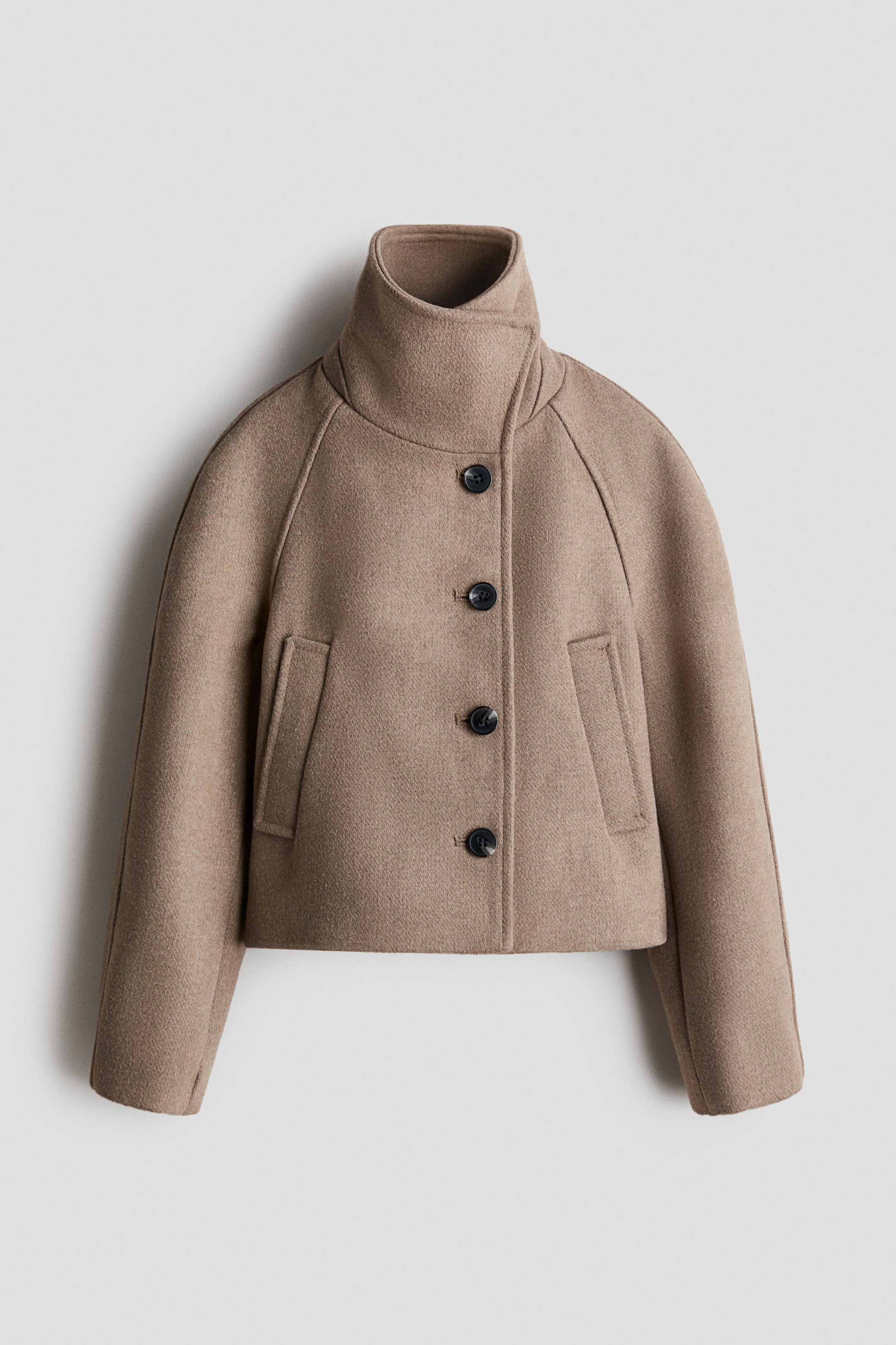 Größeres Bild anzeigen: Eine kurz geschnittene, hellbraune Wolljacke ist von vorne zu sehen und verfügt über einen hohen Stehkragen sowie einen asymmetrischen Frontverschluss mit vier dunklen Knöpfen. Die Jacke ist mit strukturierten Raglanärmeln, einer kastigen Silhouette und zwei schrägen Leistentaschen auf der Vorderseite gestaltet.