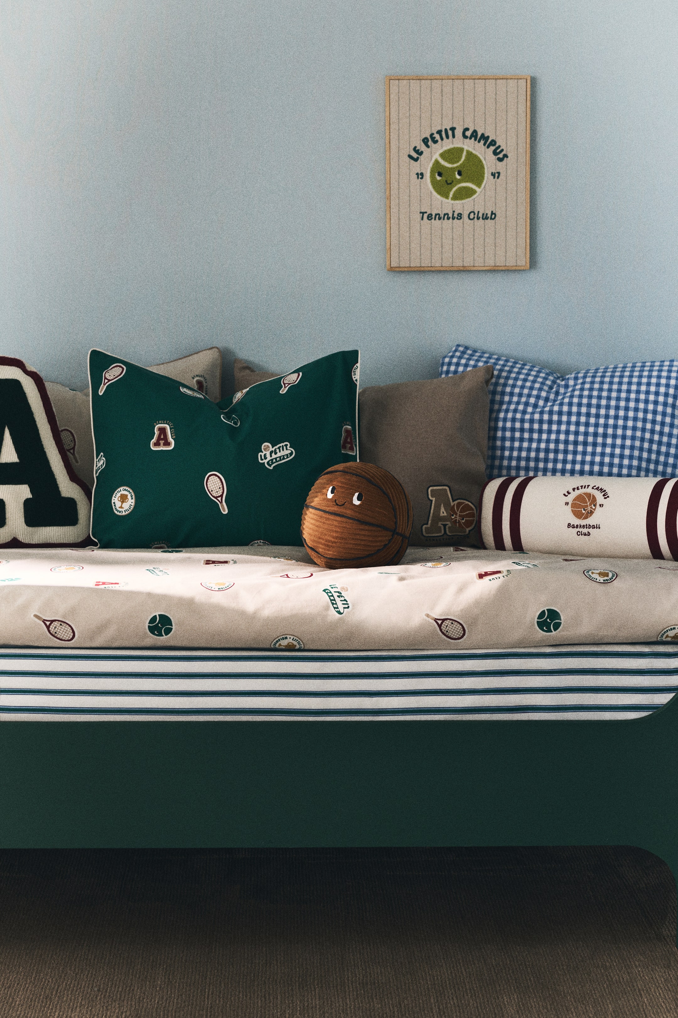 Grotere afbeelding bekijken: Een beddengoedset is gerangschikt op een donkergroen bedframe, met een beige dekbedovertrek bedrukt met tennisrackets en sportclubemblemen, gelaagd over een wit laken met groene en blauwe strepen. Verschillende kussens maken de opstelling compleet, waaronder een donkergroen kussen met tennismotieven, een bruin basketbalvormig kussen met een geborduurd gezicht, en een witte rolkussen met de tekst "Basketball Club".