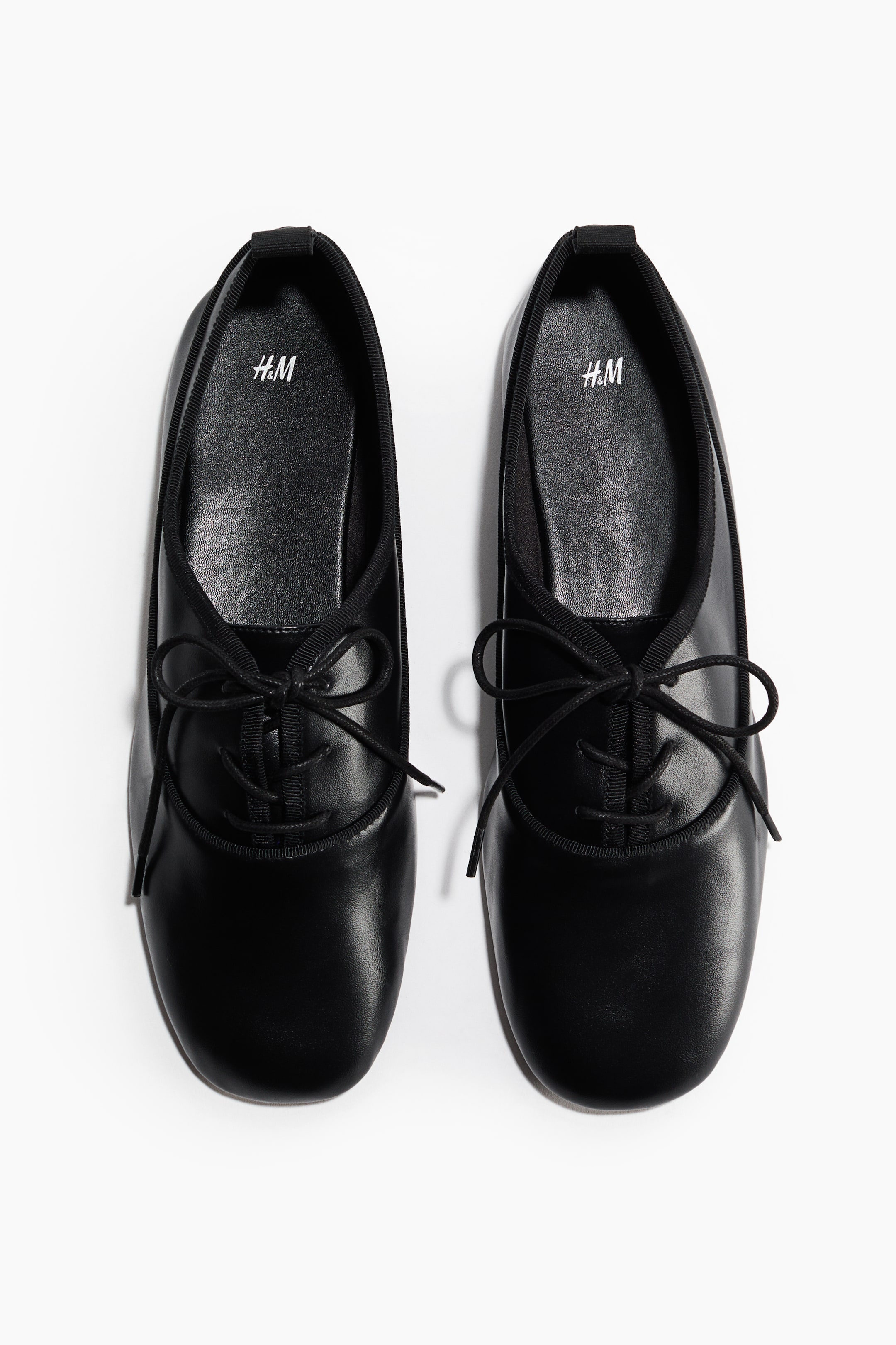 Agrandir l'image: Une paire de ballerines noires à lacets présente une tige lisse, des bouts arrondis, et de fines lacets en ruban noir noués en nœuds sur le dessus du pied. Chaque chaussure affiche également un bord côtelé noir autour de l'ouverture et un logo blanc 'H&M' sur la semelle intérieure noire.