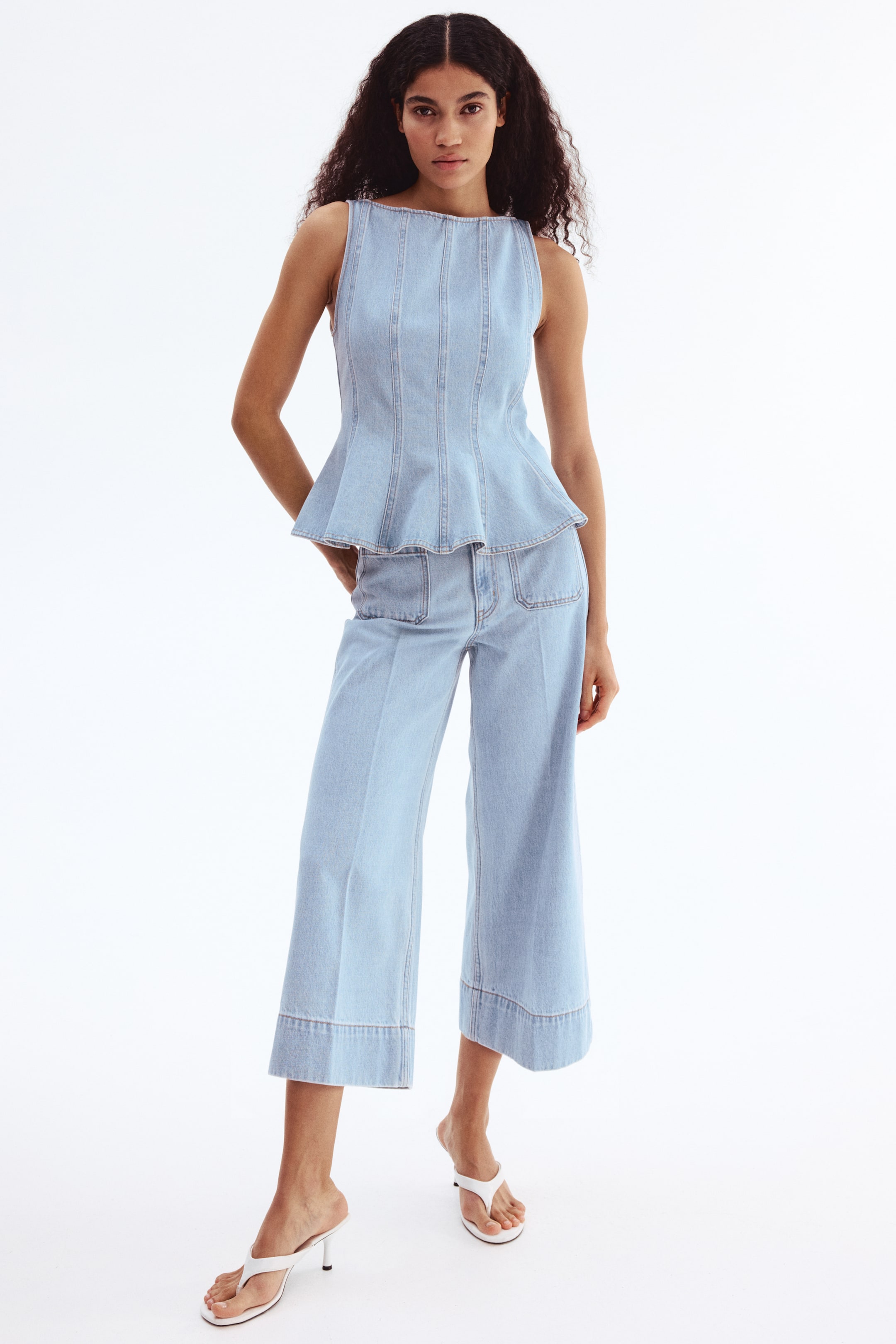 View larger image: A light blue denim sleeveless top and matching cropped wide-leg pants are showcased from a frontal perspective. The top boasts a boat neckline, vertical seaming on the bodice, and a peplum hem, while the pants are crafted with wide cuffs and front patch pockets.