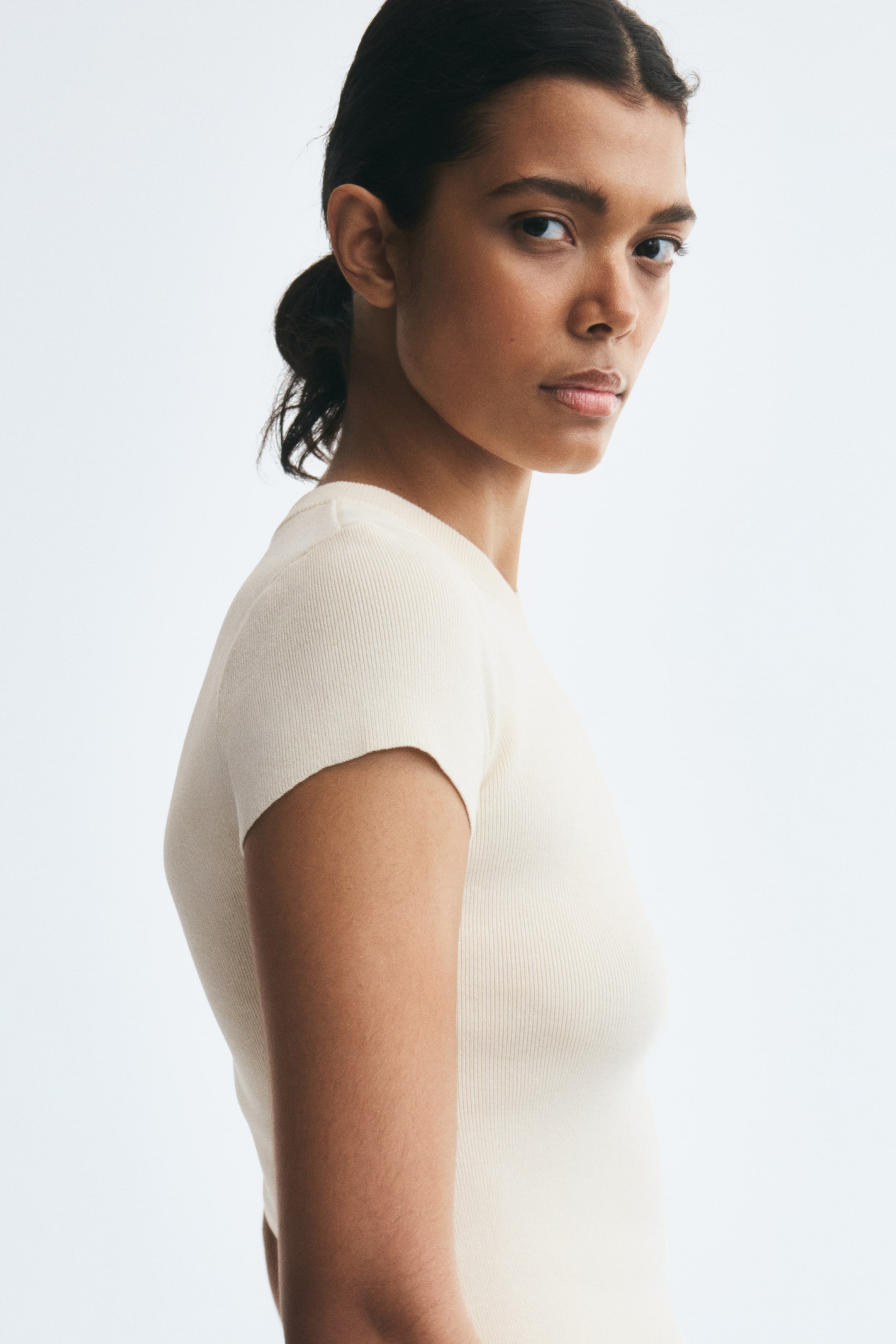 View larger image: A cream-colored, fitted ribbed-knit tee is being worn by the model in a side-profile view. The short-sleeved top features a fine ribbed texture, a crew neckline, and a snug fit.