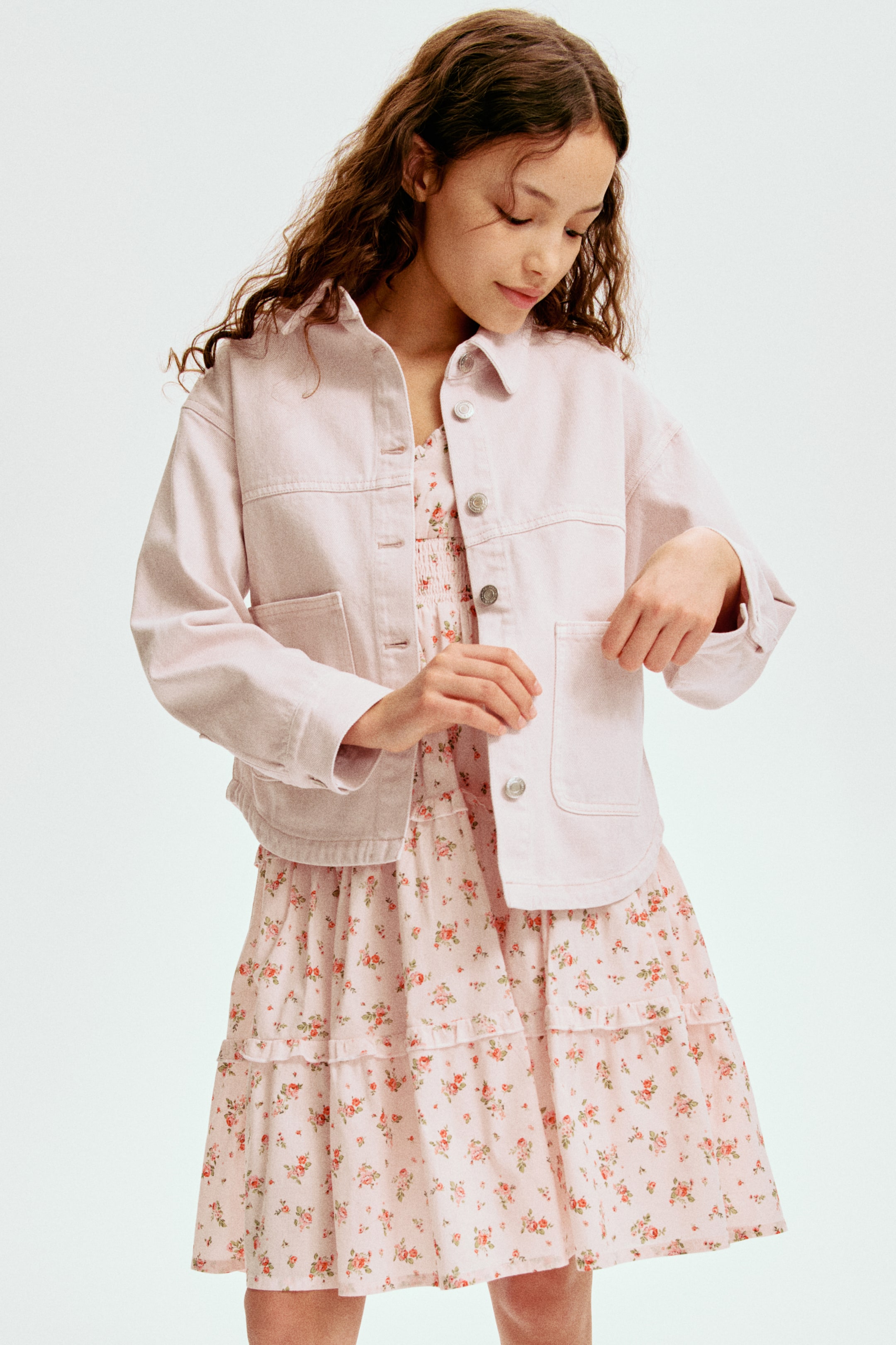 Ampliar la imagen: Una chaqueta de mezclilla rosa claro se lleva abierta sobre un vestido floral rosa claro. La chaqueta cuenta con un cuello, botones frontales en tono plateado, bolsillos en el pecho con solapas y bolsillos inferiores de parche, mientras que el vestido presenta un corpiño fruncido y una falda escalonada con detalles de volantes y pequeños estampados florales en naranja y verde.