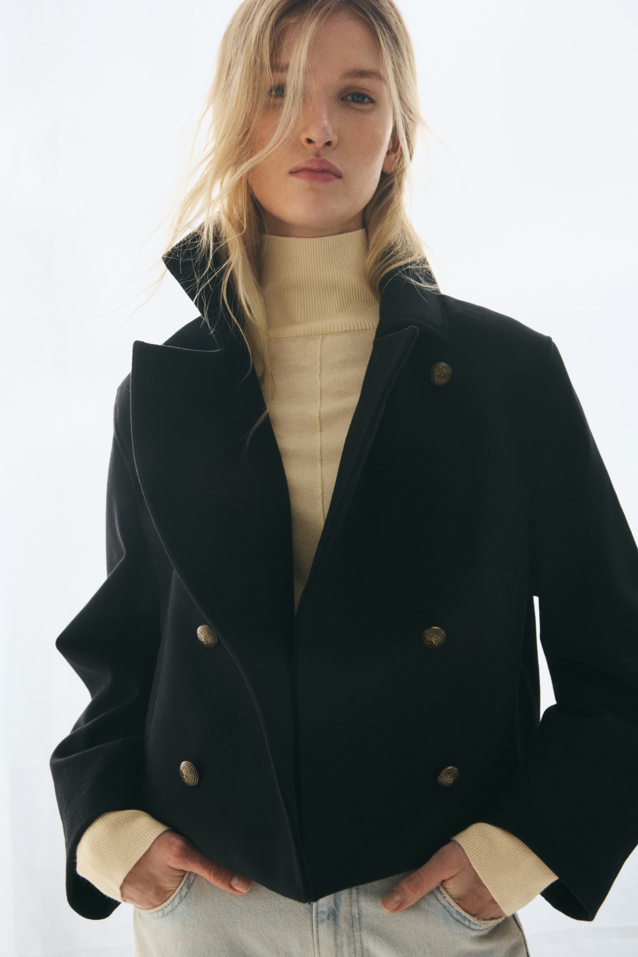 View larger image: A short, double-breasted black jacket is shown in a structured front view with a high, open funnel collar and decorative gold-toned buttons. The jacket is layered over a cream turtleneck sweater, highlighting its soft, felted texture and relaxed fit.