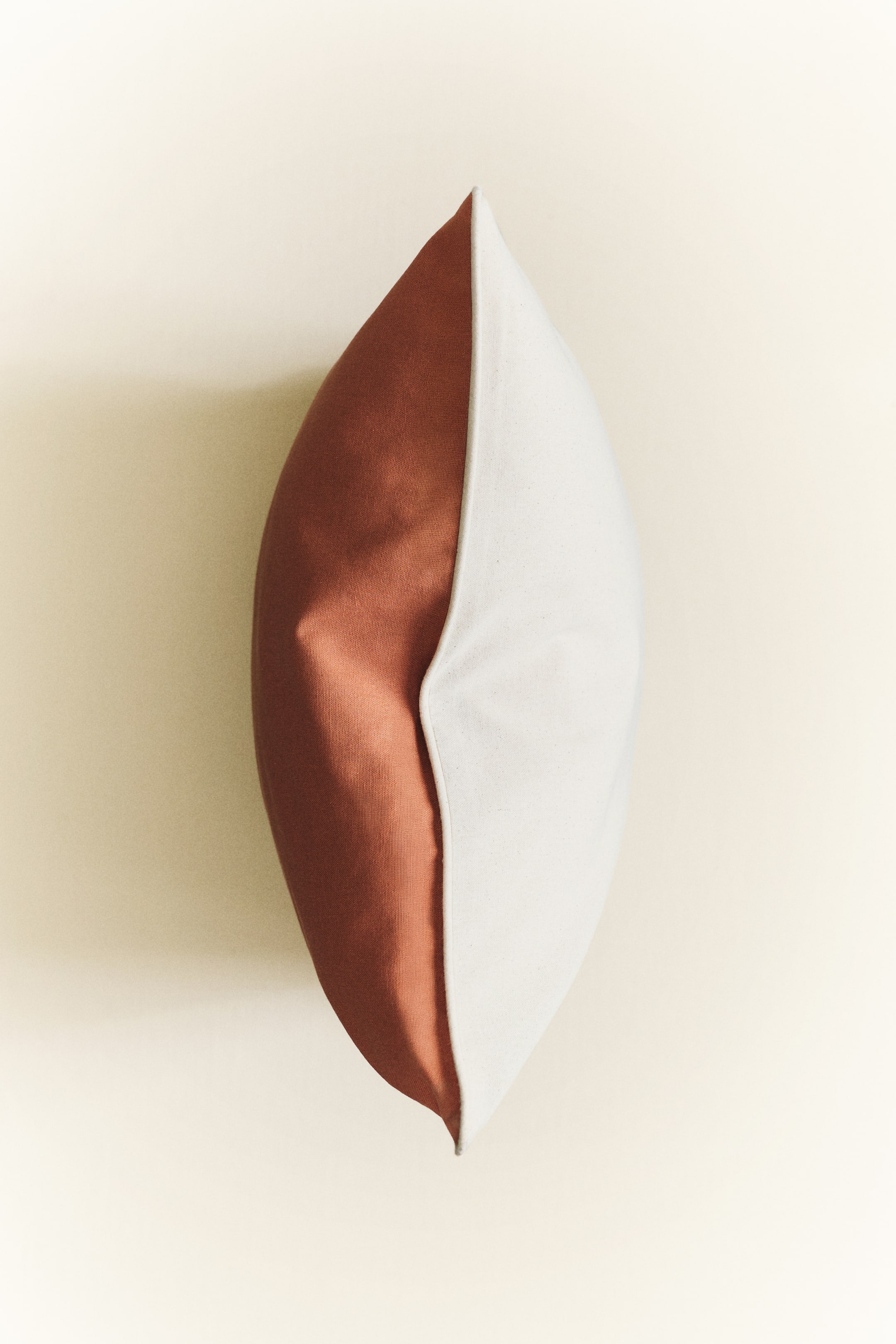 View larger image: A cotton canvas cushion cover is displayed from the side, highlighting its two-toned design with a terracotta hue on one half and an off-white shade on the other. Slim piping runs along the seam where the two colours converge.