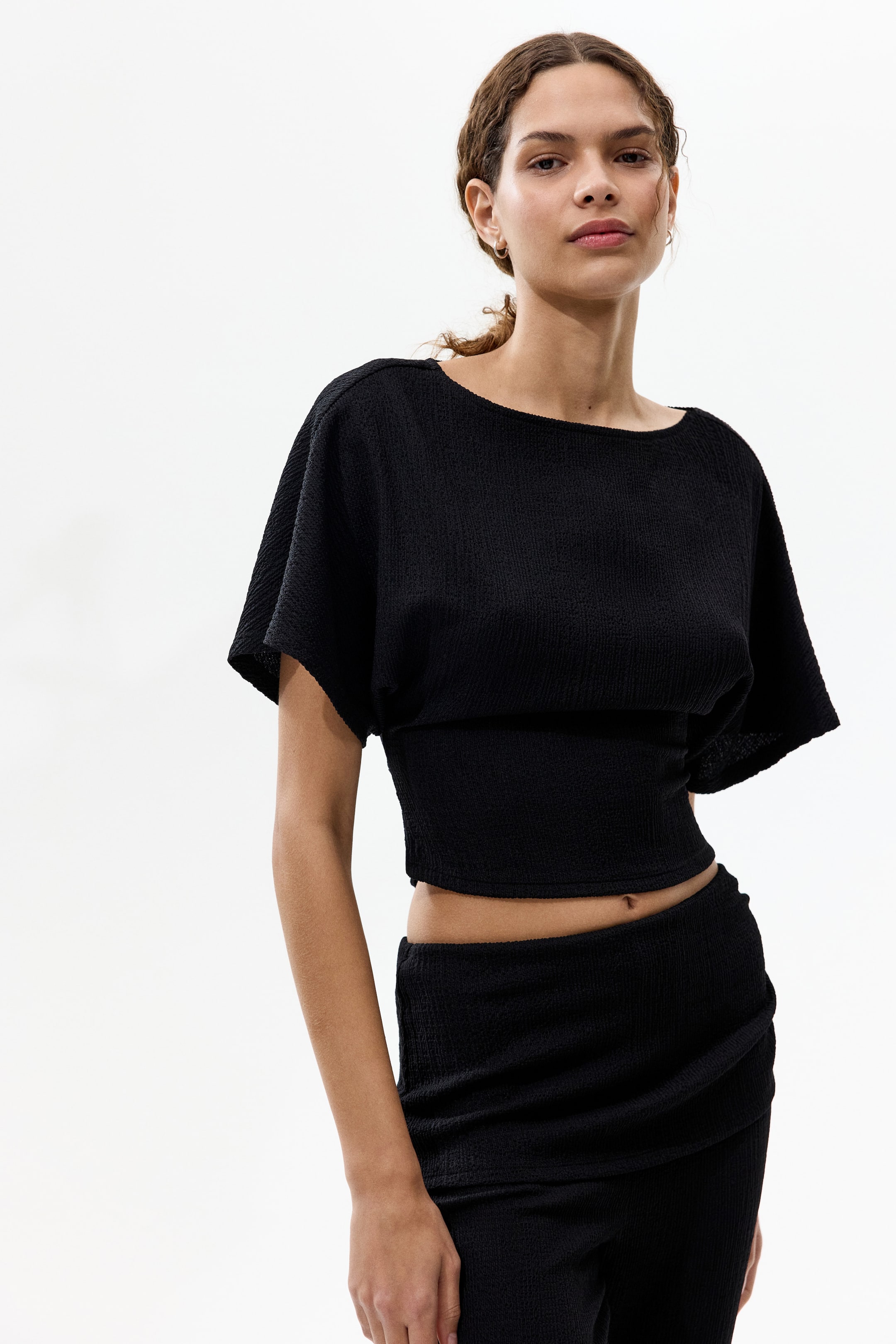 View larger image: A black textured cropped top features a boat neckline and dolman sleeves, ending just above the navel. It is paired with a matching black textured skirt, which showcases a draped front detail and sits low on the hips.