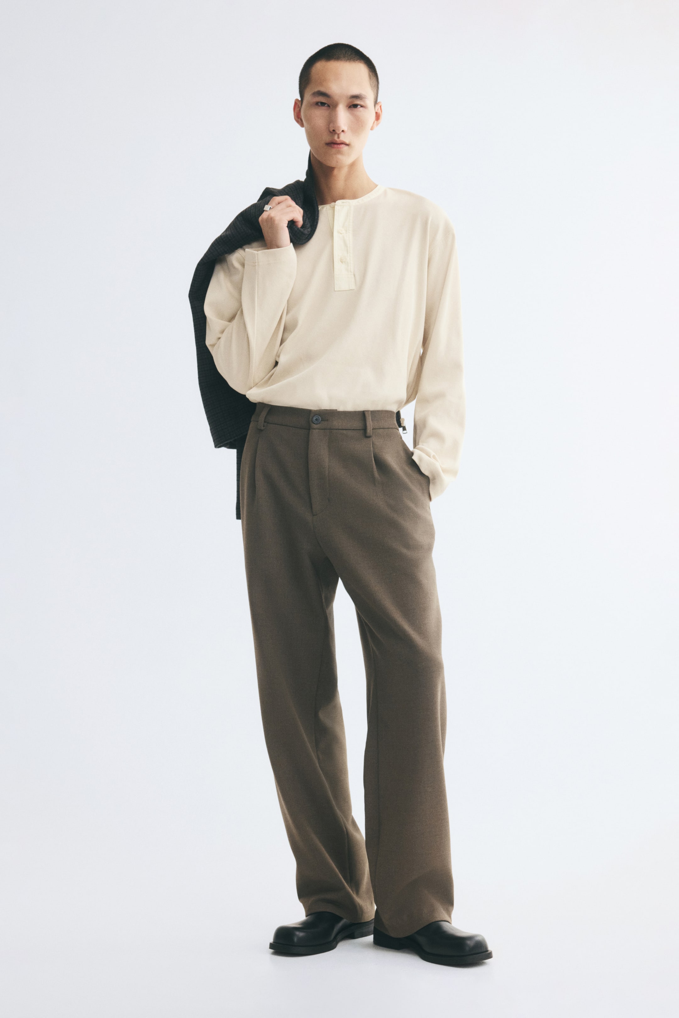 View larger image: Tailored trousers in brown woven fabric are worn by the model, featuring a relaxed fit, straight legs with front pleats, and a button-closure waist. The model is standing with one hand in a diagonal side pocket, styling the trousers with a tucked-in cream long-sleeve shirt and holding a dark jacket over one shoulder.