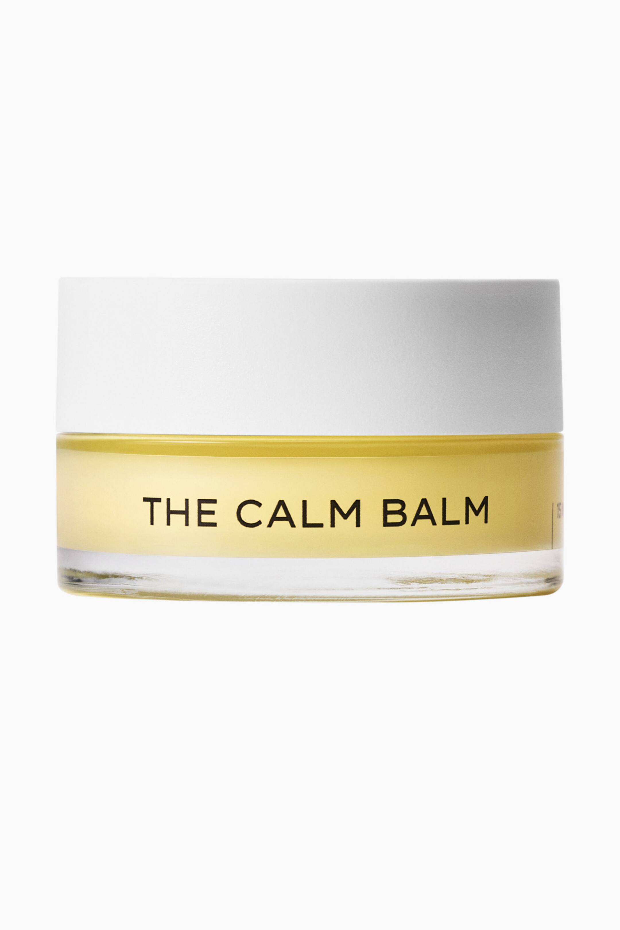 Visa större bild: En klar glasburk med "The Calm Balm" är vänd framåt och innehåller en gulaktig balm. Ett vitt lock täcker burken, och orden "THE CALM BALM" är tryckta i svart på framsidan.
