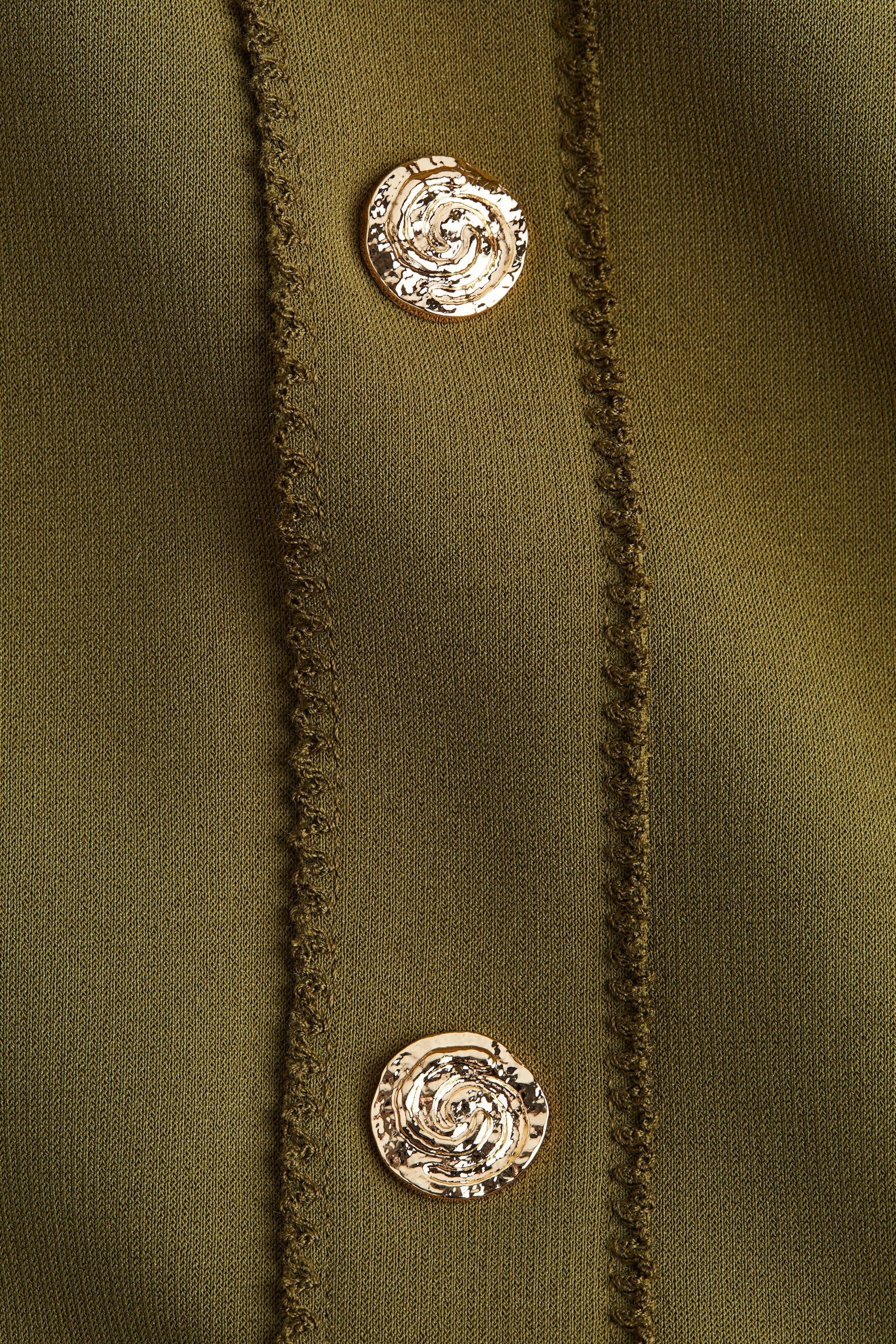 Büyük resmi görüntüle: Zeytin yeşili ağır interlok jarse kumaştan yapılmış dar kesim bir yelek, ön kısmında çekiçle işlenmiş spiral tasarıma sahip iki dekoratif, altın tonlu yuvarlak düğme sergiliyor. Bu düğmeler, iki paralel yükseltilmiş, dokulu dikiş hattı ile çerçeveleniyor.