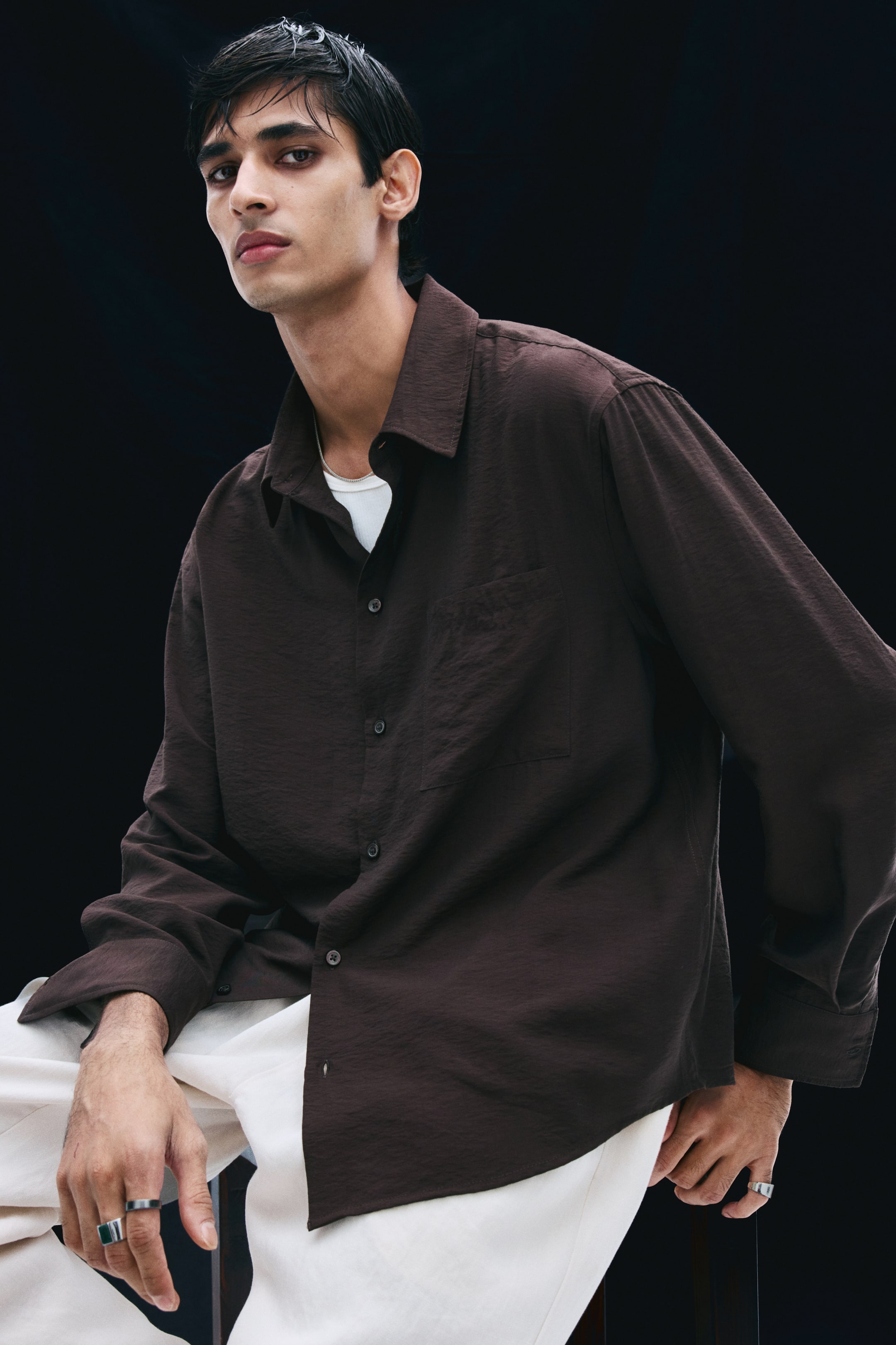 View larger image: A dark brown shirt in a lightly crinkled fabric is being worn by the model, featuring a relaxed fit, a collar, a button-up front, and a single patch chest pocket. The shirt is styled open at the collar, layered over a white t-shirt, and paired with white trousers.