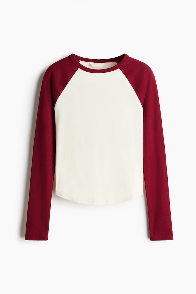 A fitted cotton jersey top is shown from the front, featuring an off-white body with a curved hem, contrasting burgundy long raglan sleeves, and a matching burgundy round neckline. - 1