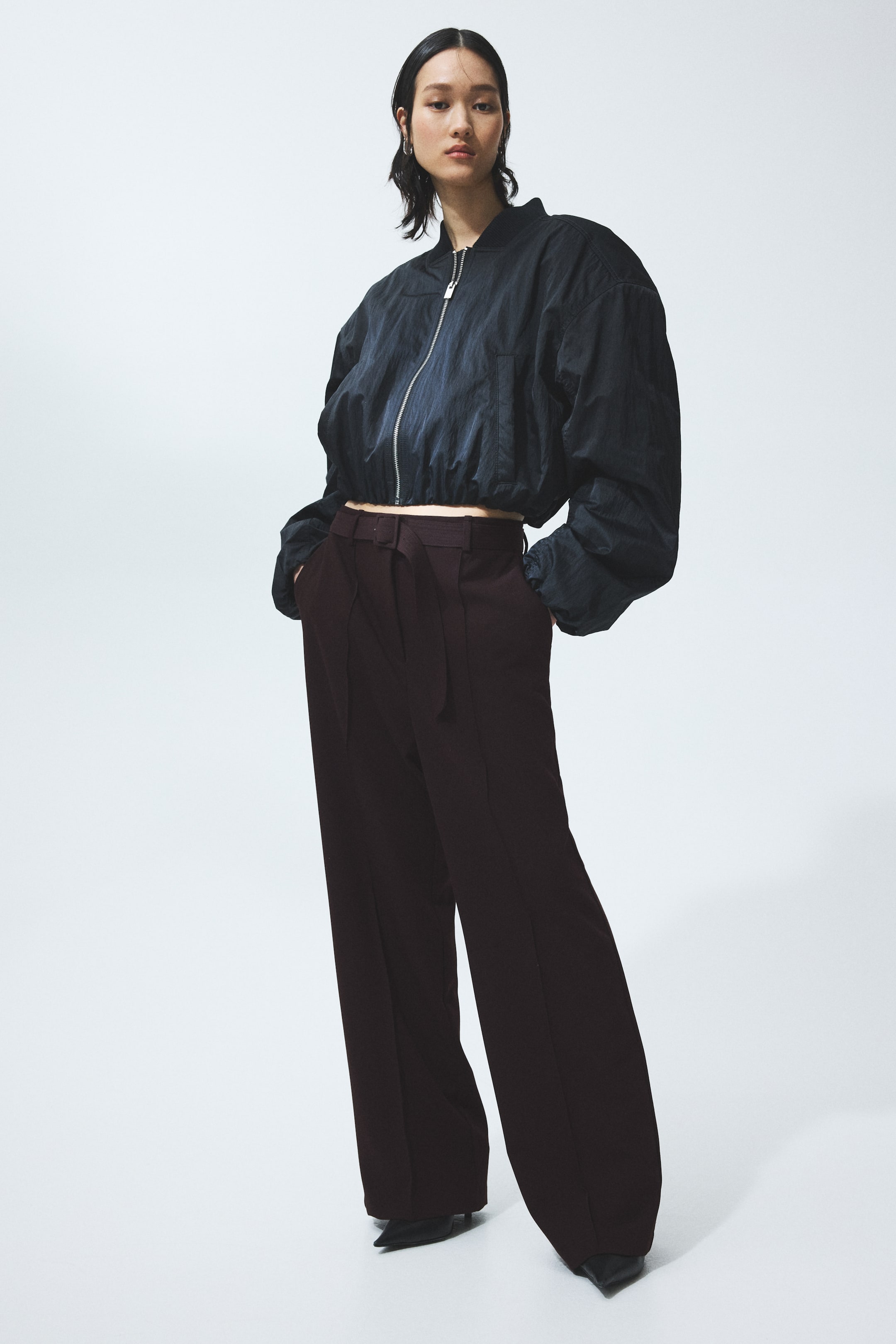 View larger image: A dark navy cropped bomber jacket features a full-zip front, a stand-up collar, and voluminous sleeves with an elasticized hem. It layers over dark brown high-waisted wide-leg trousers, which display belt loops and subtle vertical seam details, partially covering pointed-toe shoes.