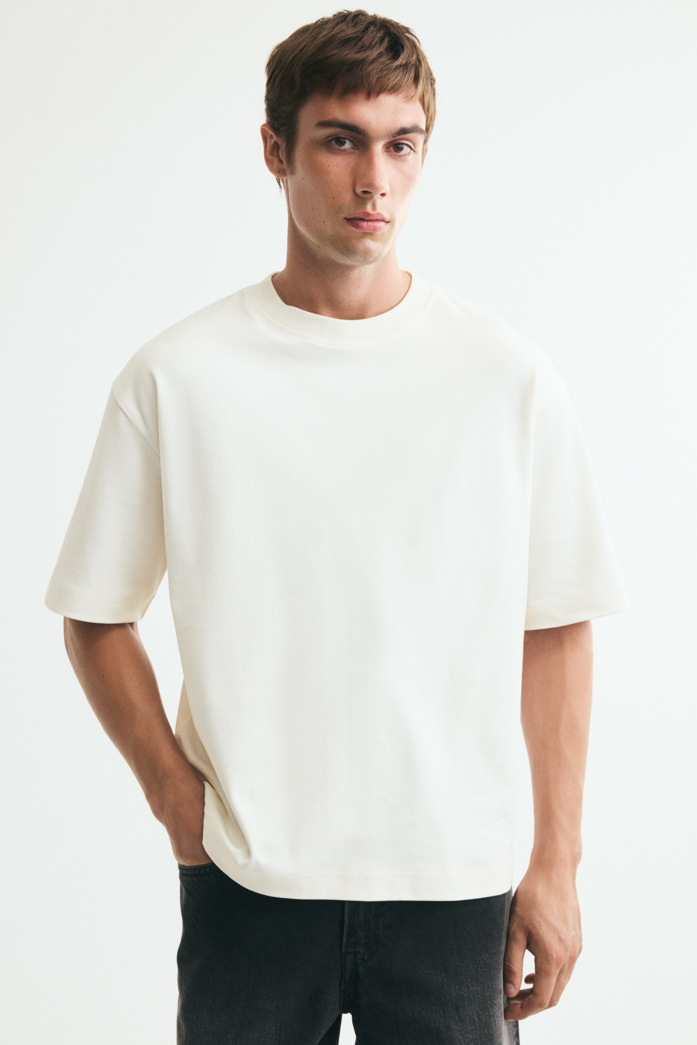 Uma t-shirt creme exibe a sua frente, apresentando uma gola redonda canelada com uma etiqueta preta da marca H&M e mangas curtas, oferecendo um corte descontraído.