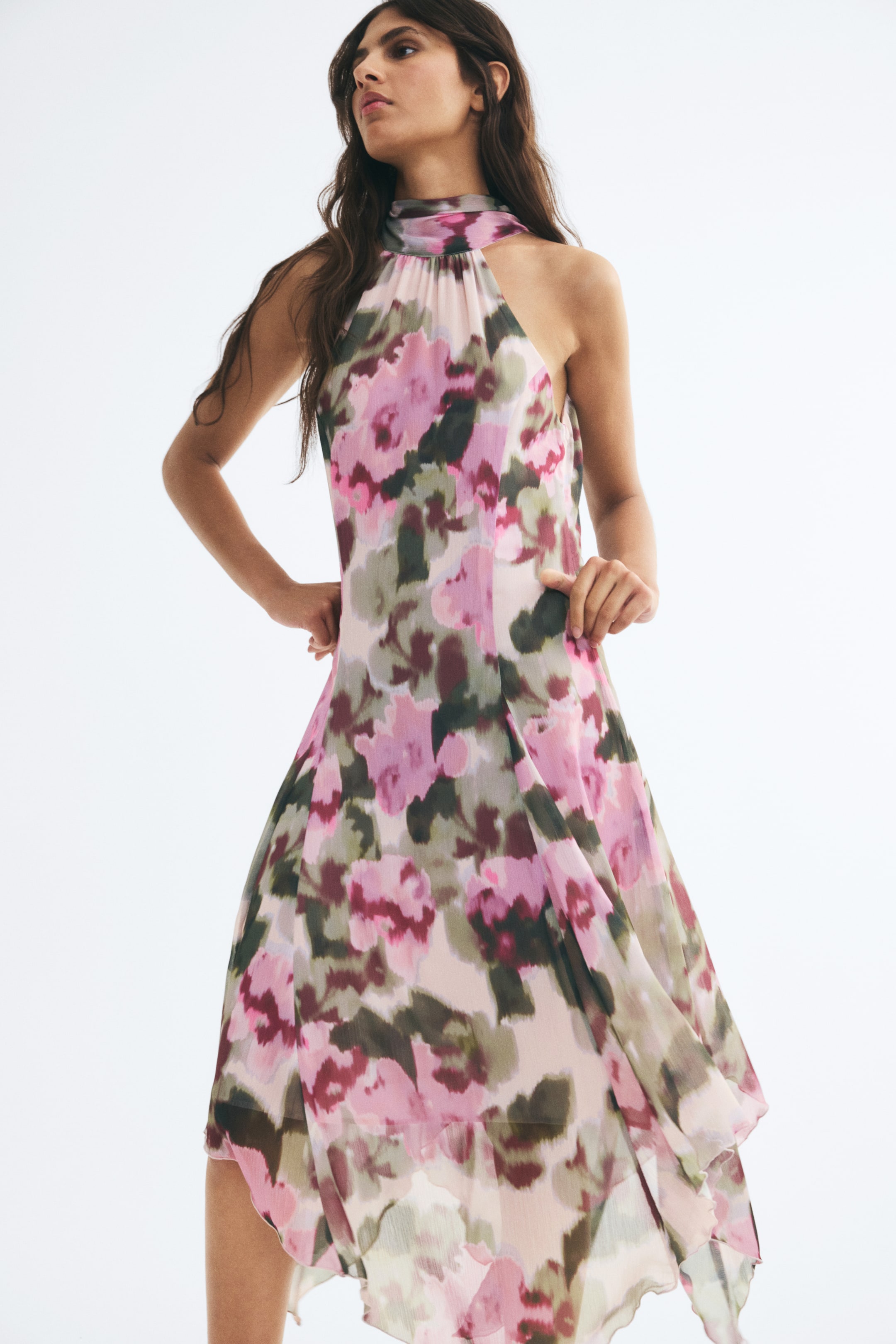 View larger image: The model is wearing a long, sleeveless dress in crinkled chiffon with a pink, purple, and green floral print. The dress features a high gathered collar and a flowing A-line skirt with an asymmetrical handkerchief hem.