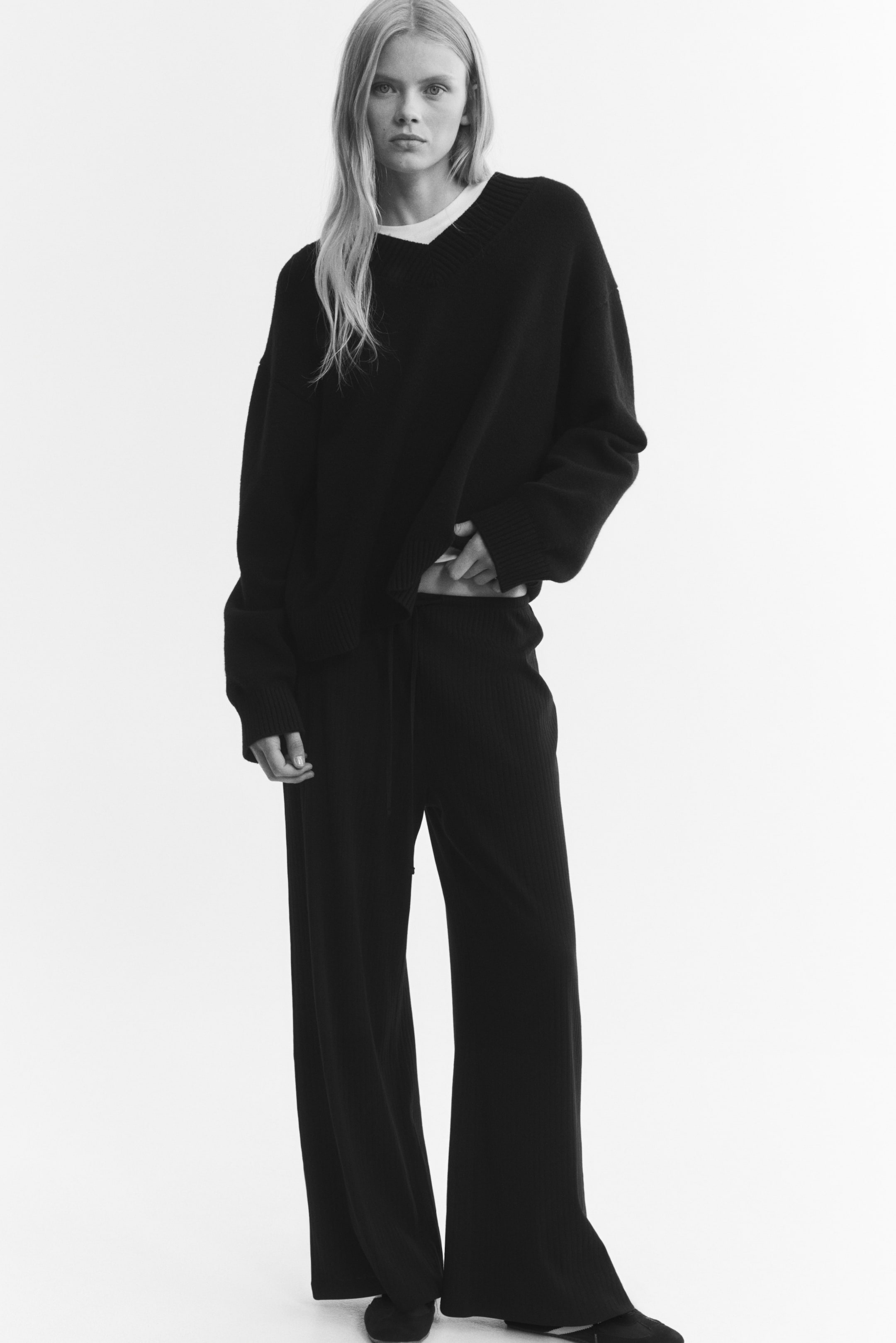 View larger image: Loose-fit pants in ribbed cotton jersey are worn by the model, featuring a wide-leg cut and an elasticised drawstring waist. The model is lifting the hem of a matching dark knit sweater to partially reveal the waistband detail.
